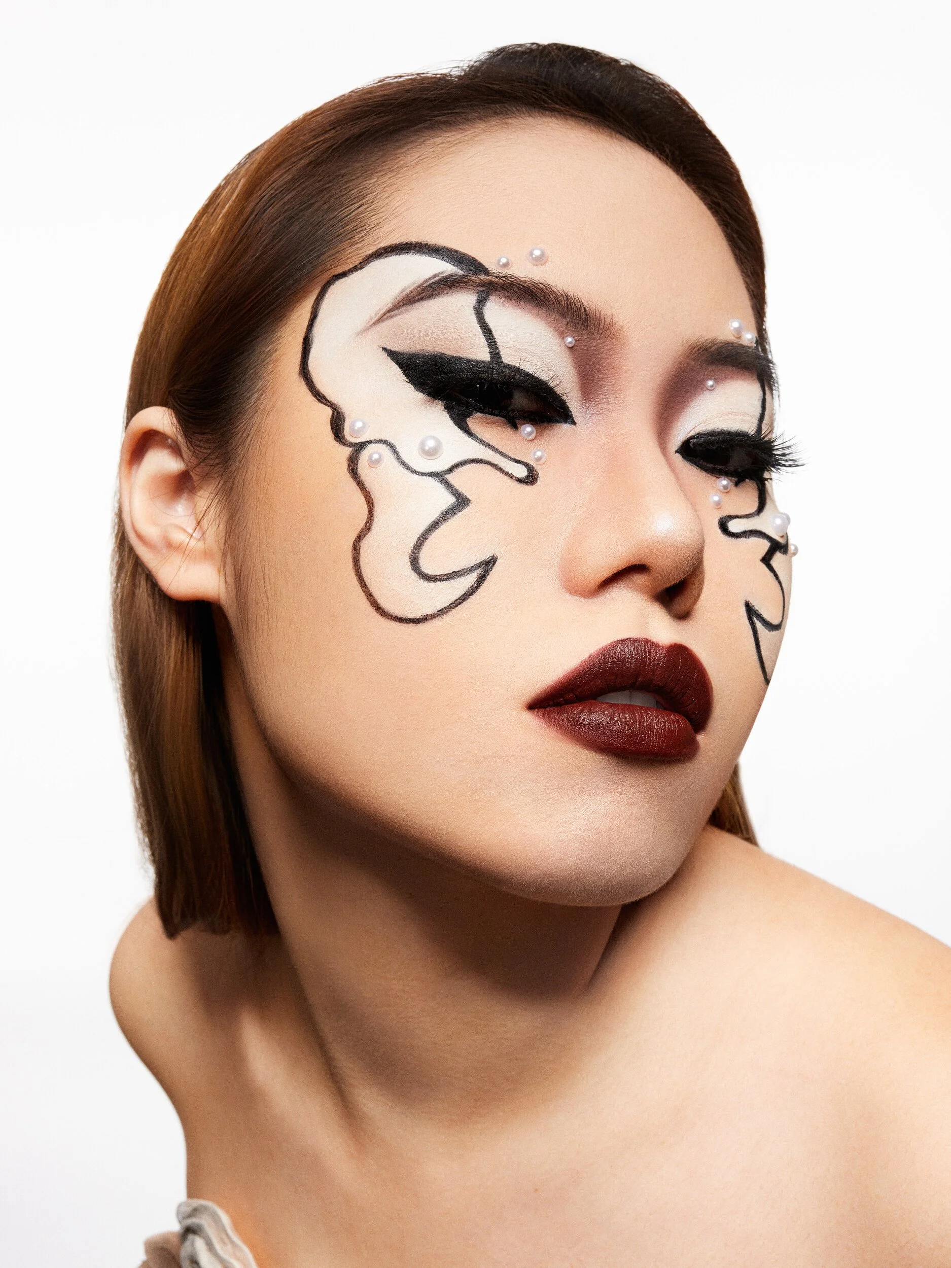 Beauty portrait with graphic black liner swirls and pearl accents around the eyes and deep burgundy lips on a white backdrop