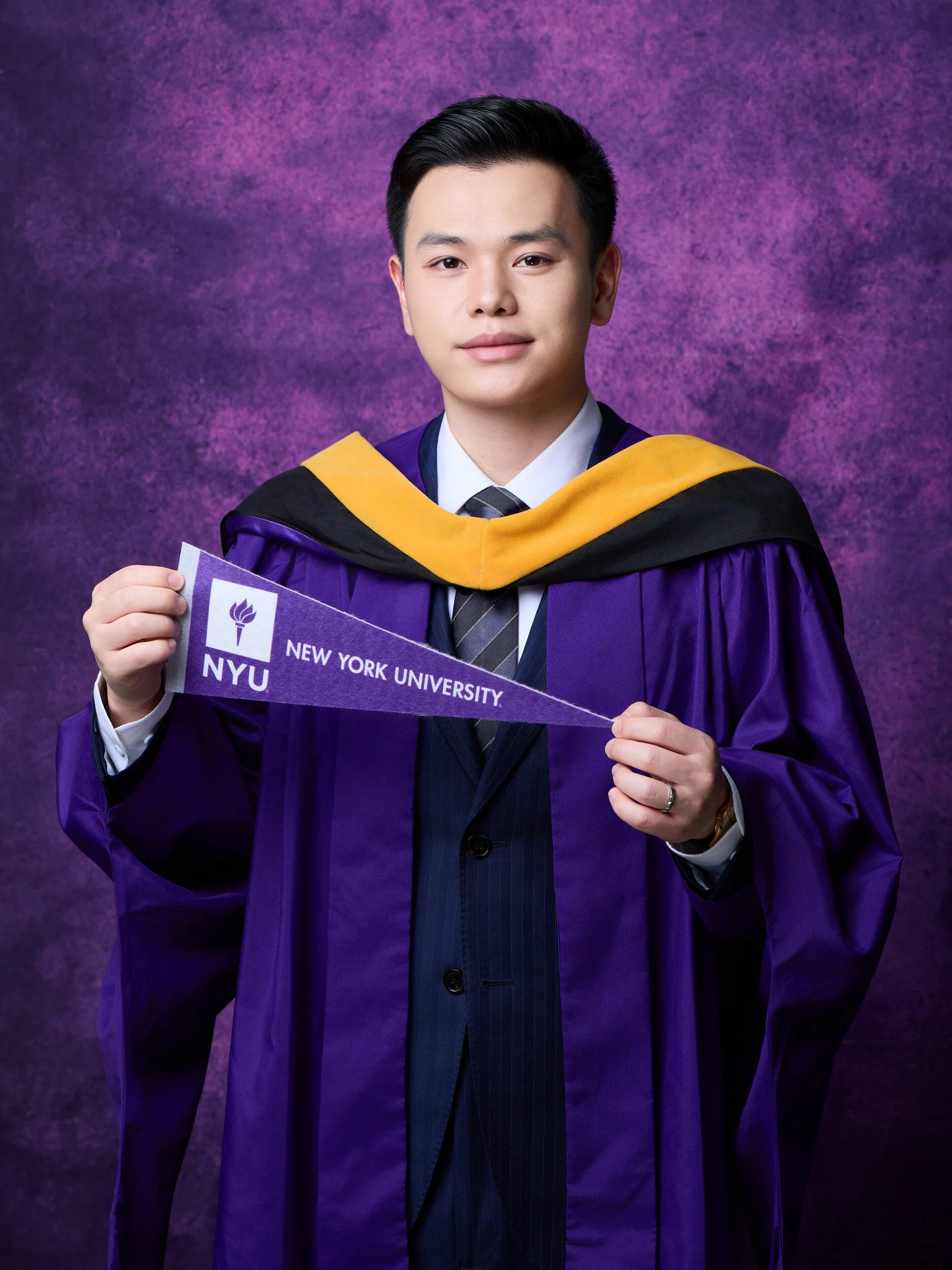 NYU graduation portrait of a young man in a navy suit under his gown holding a university pennant