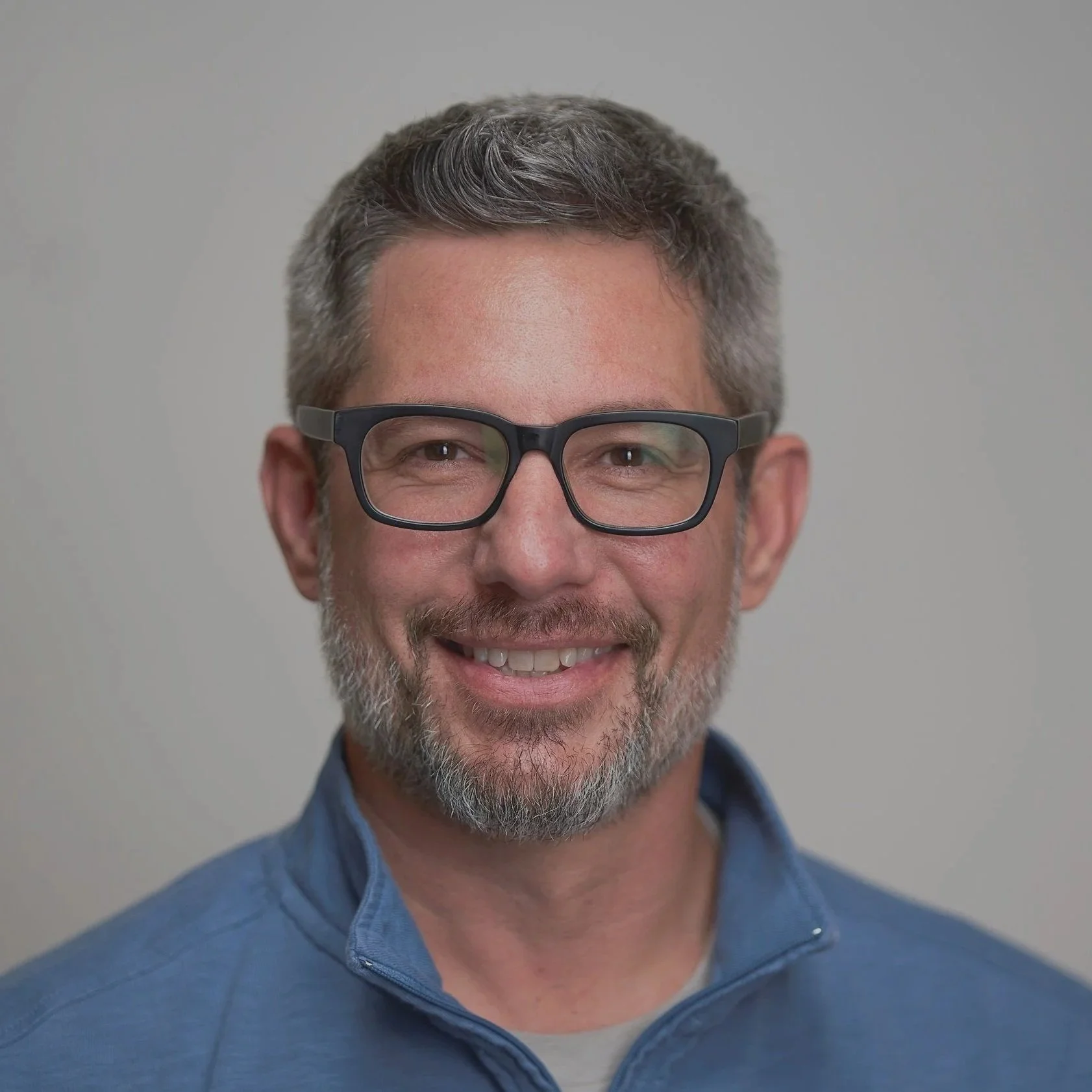A middle-aged man with gray hair and beard, wearing black-rimmed glasses and a blue jacket, smiling at the camera against a plain light gray background.