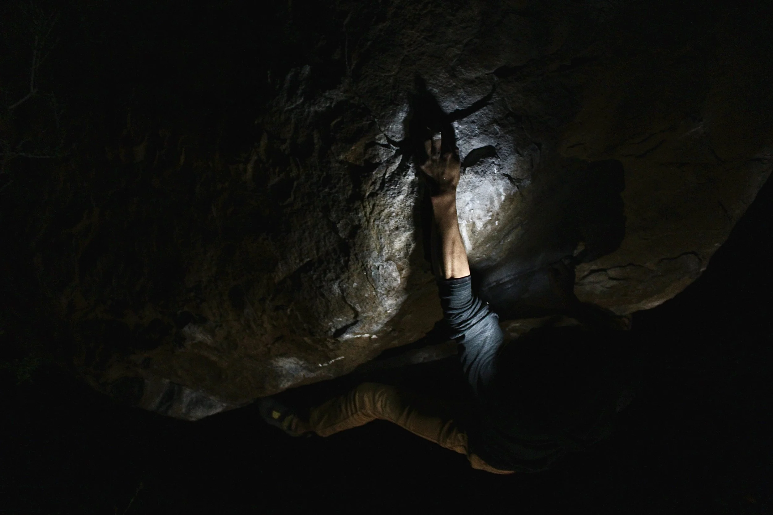 A person is bouldering on a dark rock face in a cave-like setting, with only their arm and part of their leg visible, illuminated by a flashlight or headlamp.