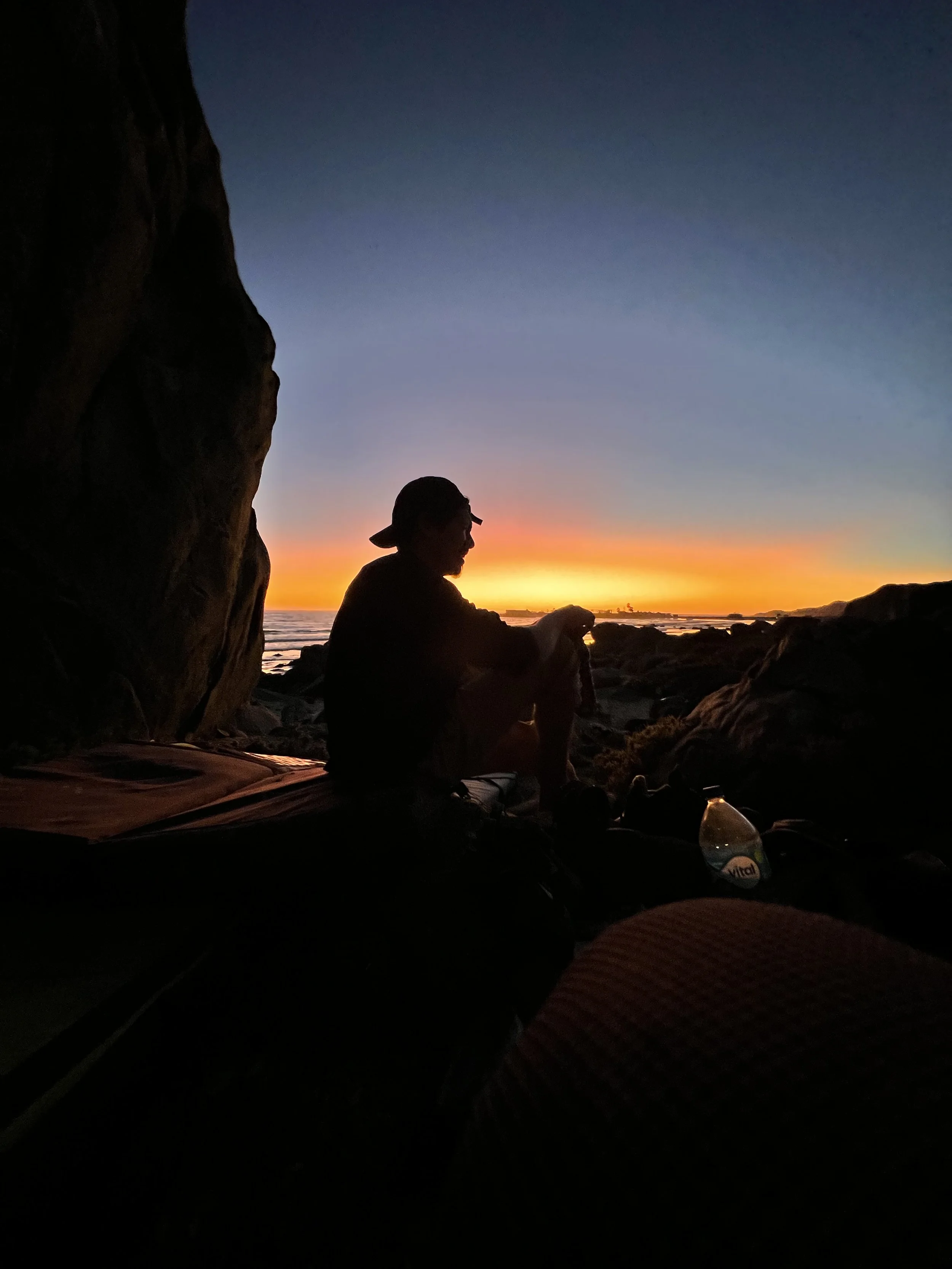 A person sitting inside a rocky cave during sunset, silhouetted against the colorful sky with the ocean in the background.