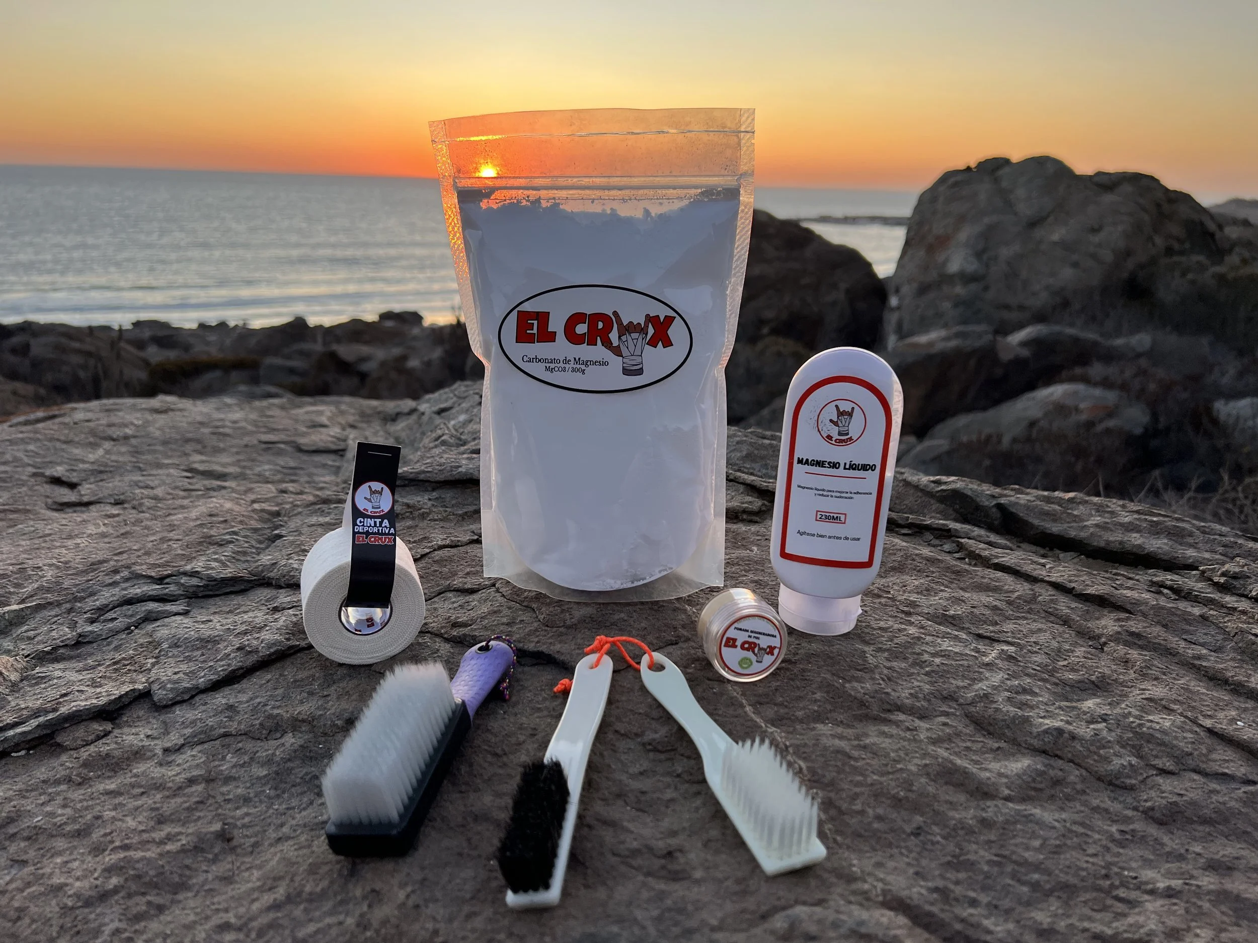Collection of climbing accessories on a rocky surface at sunset, including a bag of magnesium carbonate, a bottle of liquid magnesium, a roll of athletic tape, a small container, and bouldering and climbing brushes.