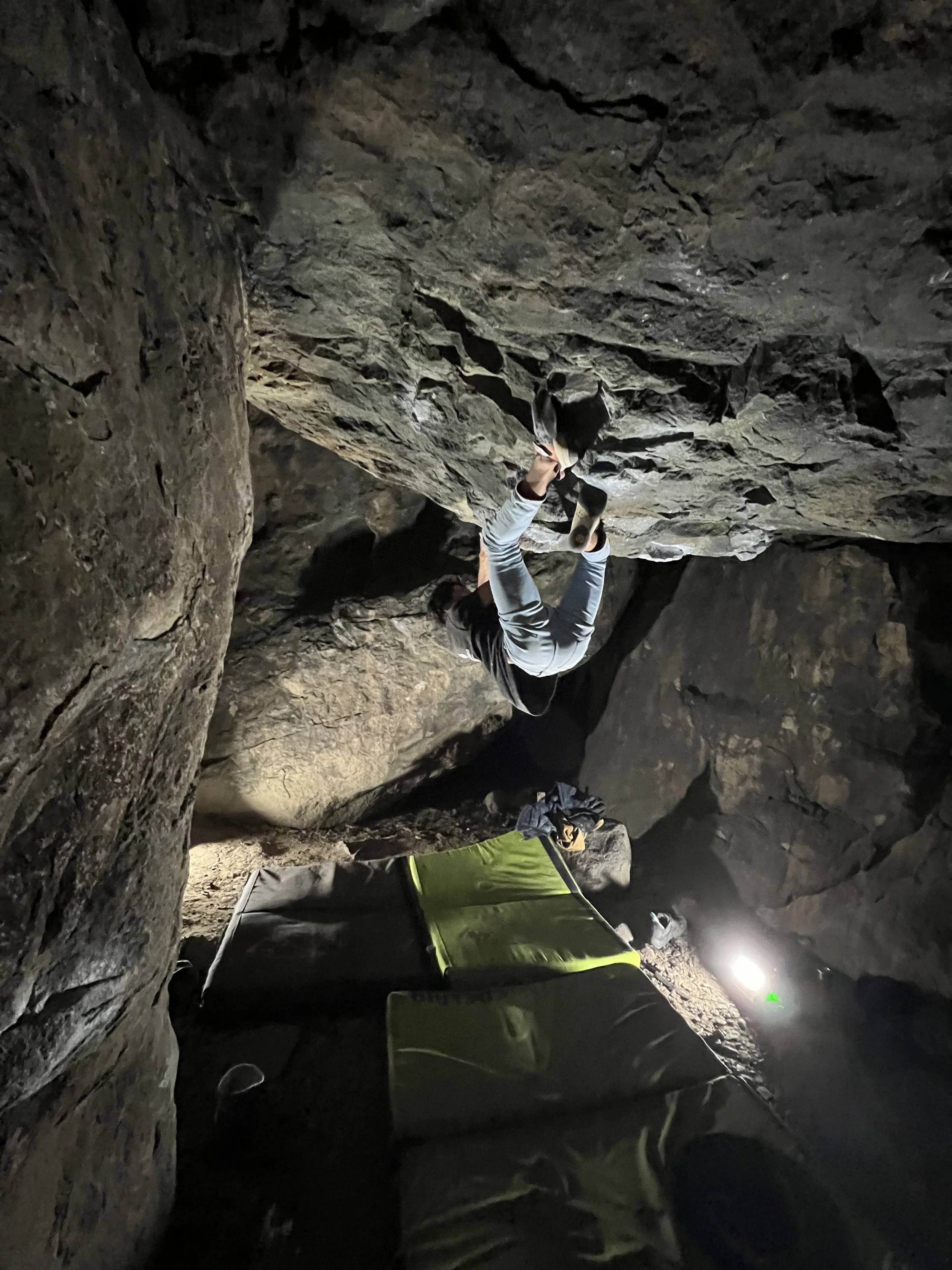 Person in a gray shirt and pants rock climbing in a dark cave, using advanced climbing shoes, with a backpack and gear on the ground nearby and a bright light illuminating the cave interior.