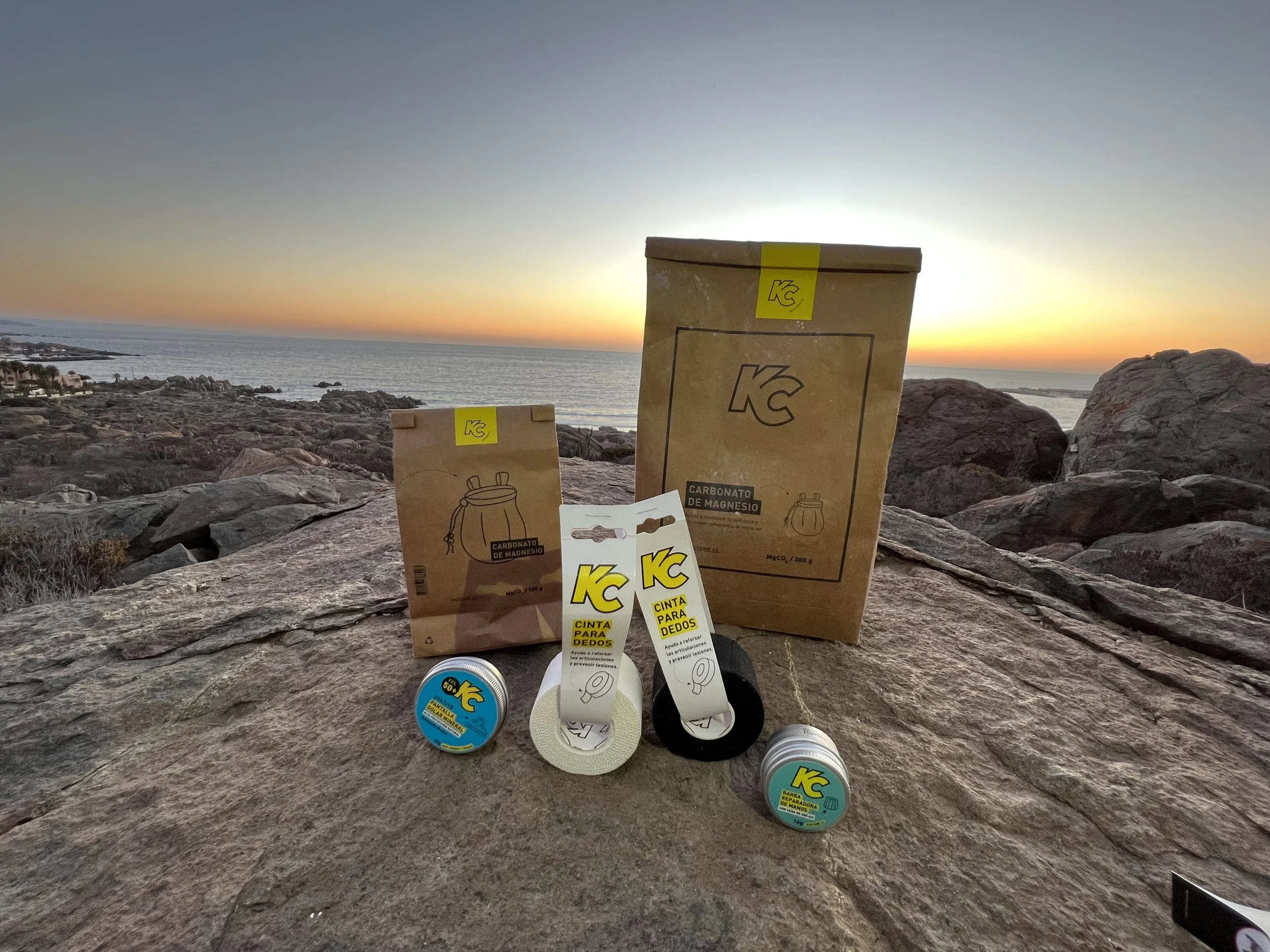 A collection of KC brand magnesium carbonate products, including boxes, tins, and finger tapes, displayed on a flat rocky surface outdoors with a sunset over the ocean in the background.