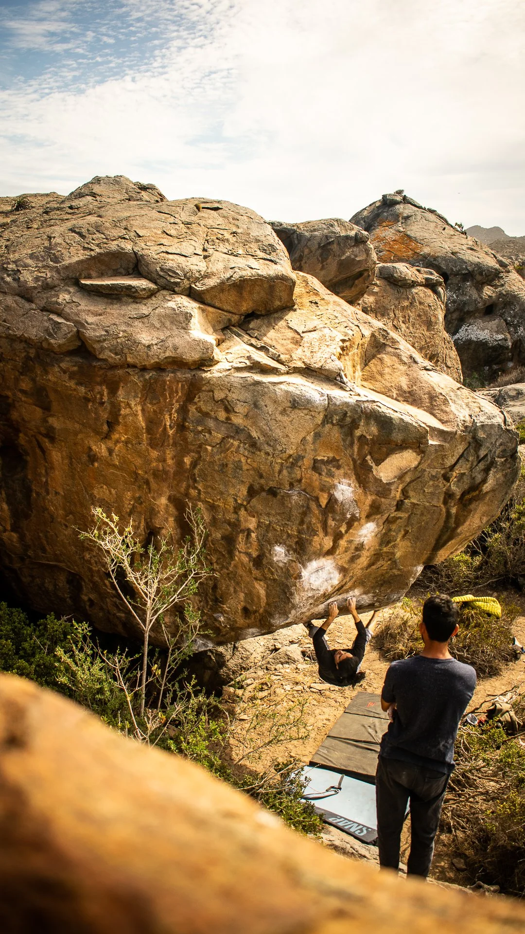 A person bouldering on a large rock over a crash pad, with another person standing nearby, outdoors in a natural rocky and shrub environment under a partly cloudy sky.