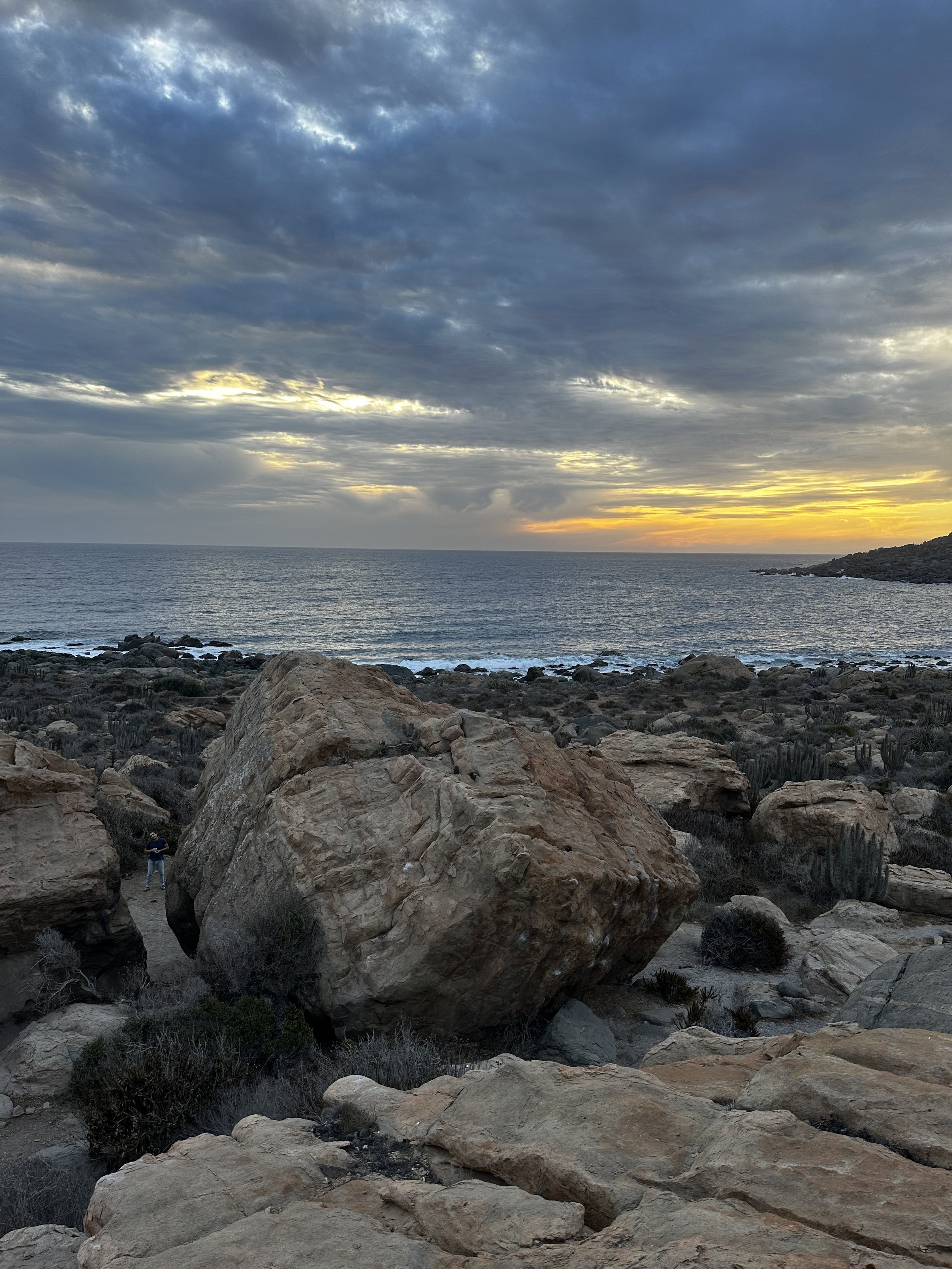 A rocky coastline at sunset, with large boulders in the foreground, ocean waves in the middle, and a partly cloudy sky with the sun setting on the horizon.