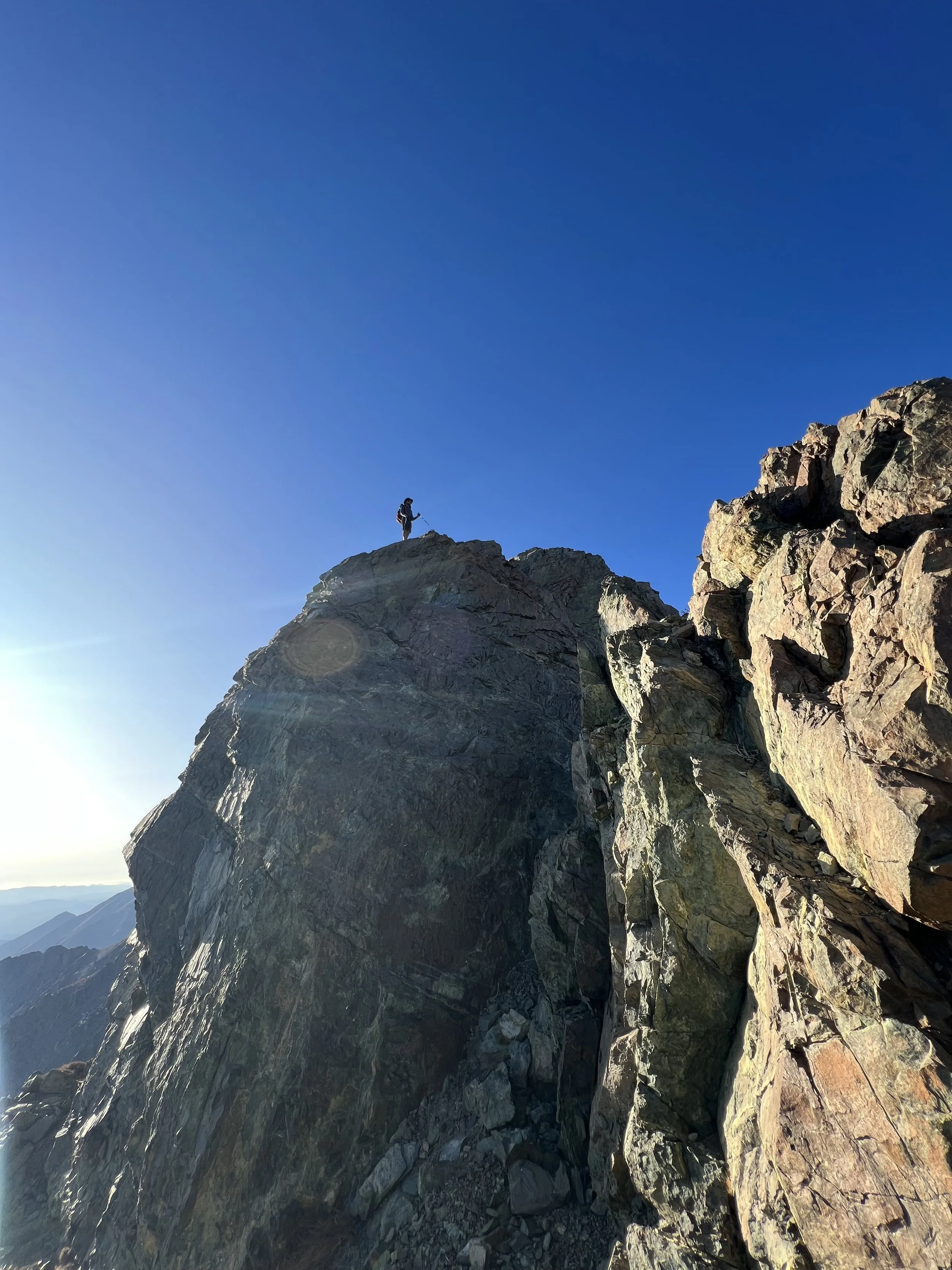 A hiker standing on the edge of a rocky mountain peak against a clear blue sky during daylight.
