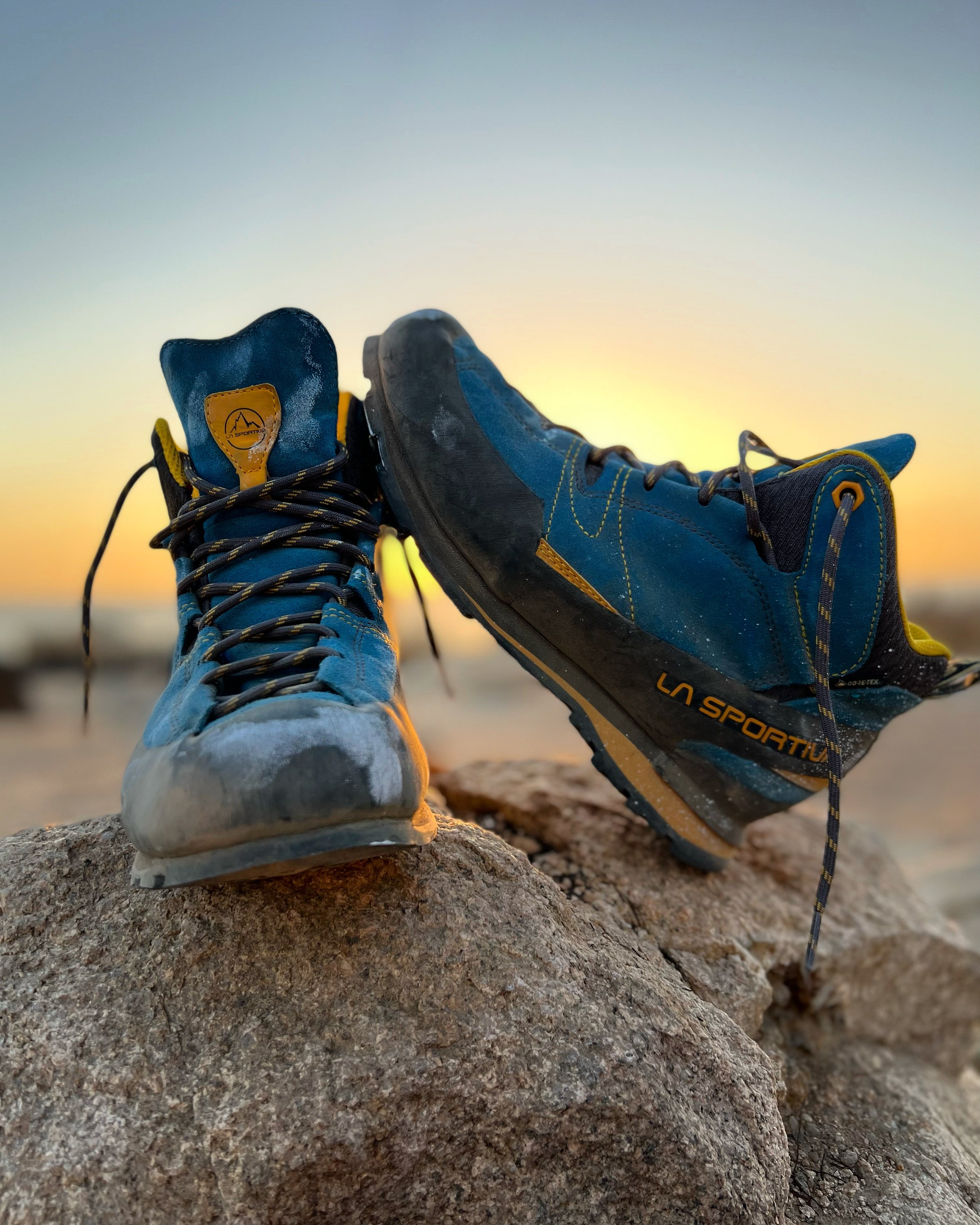Pair of blue La Sportiva outdoor hiking shoes on rocks at sunset, with a blurred background of the sky and horizon.