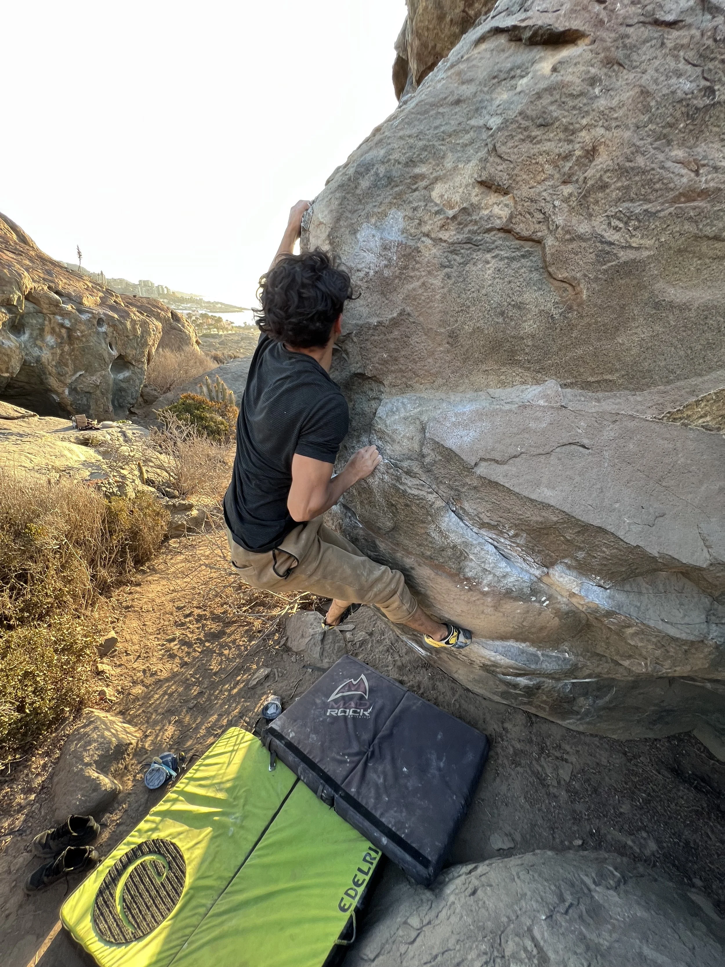 A man is rock climbing on a large outdoor boulder during sunset, with climbing gear and crash pads on the ground below.