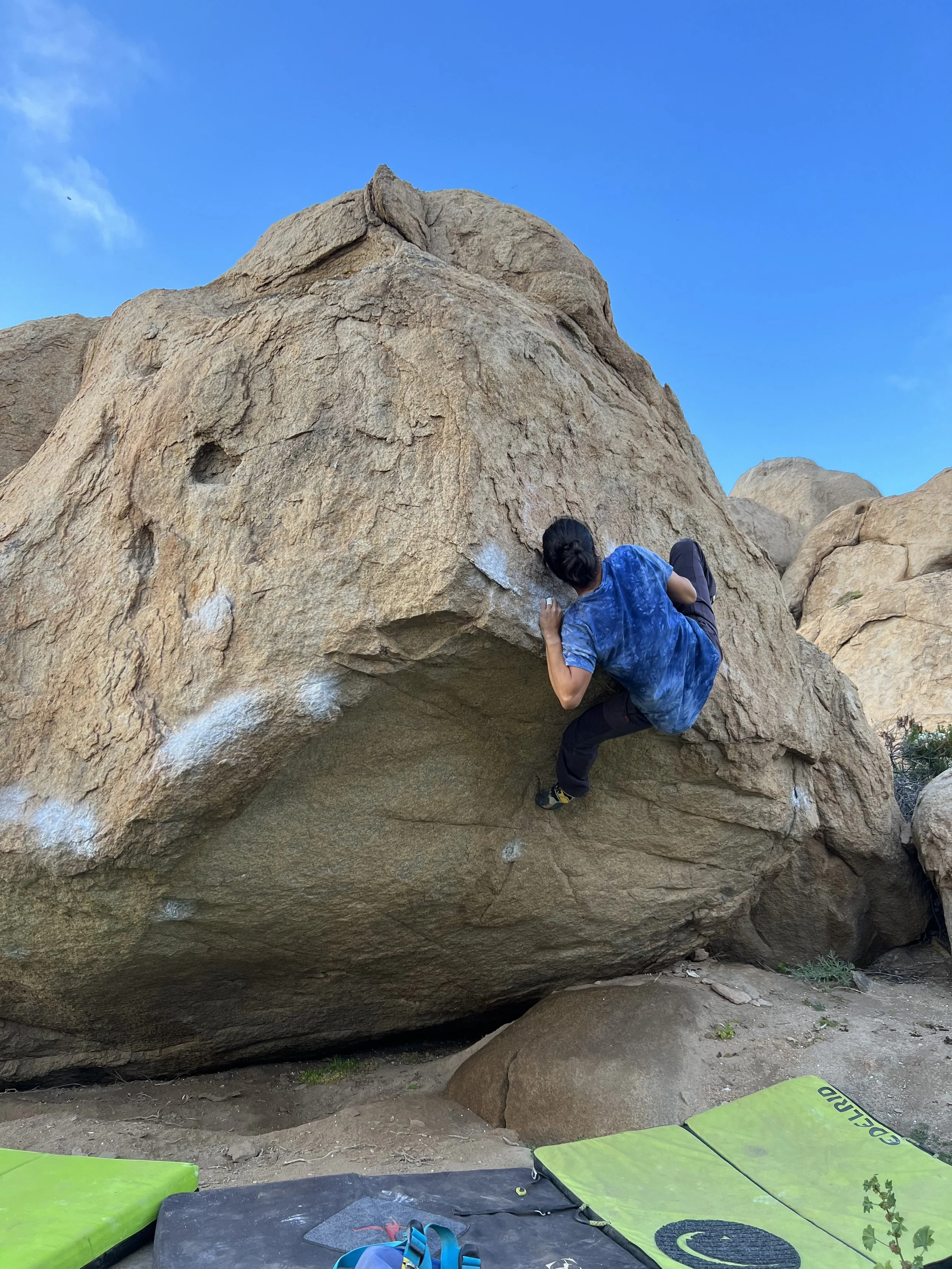 A person bouldering on a large outdoor rock, with chalk marks indicating climbing routes, and crash pads placed on the ground for safety, under a clear blue sky.