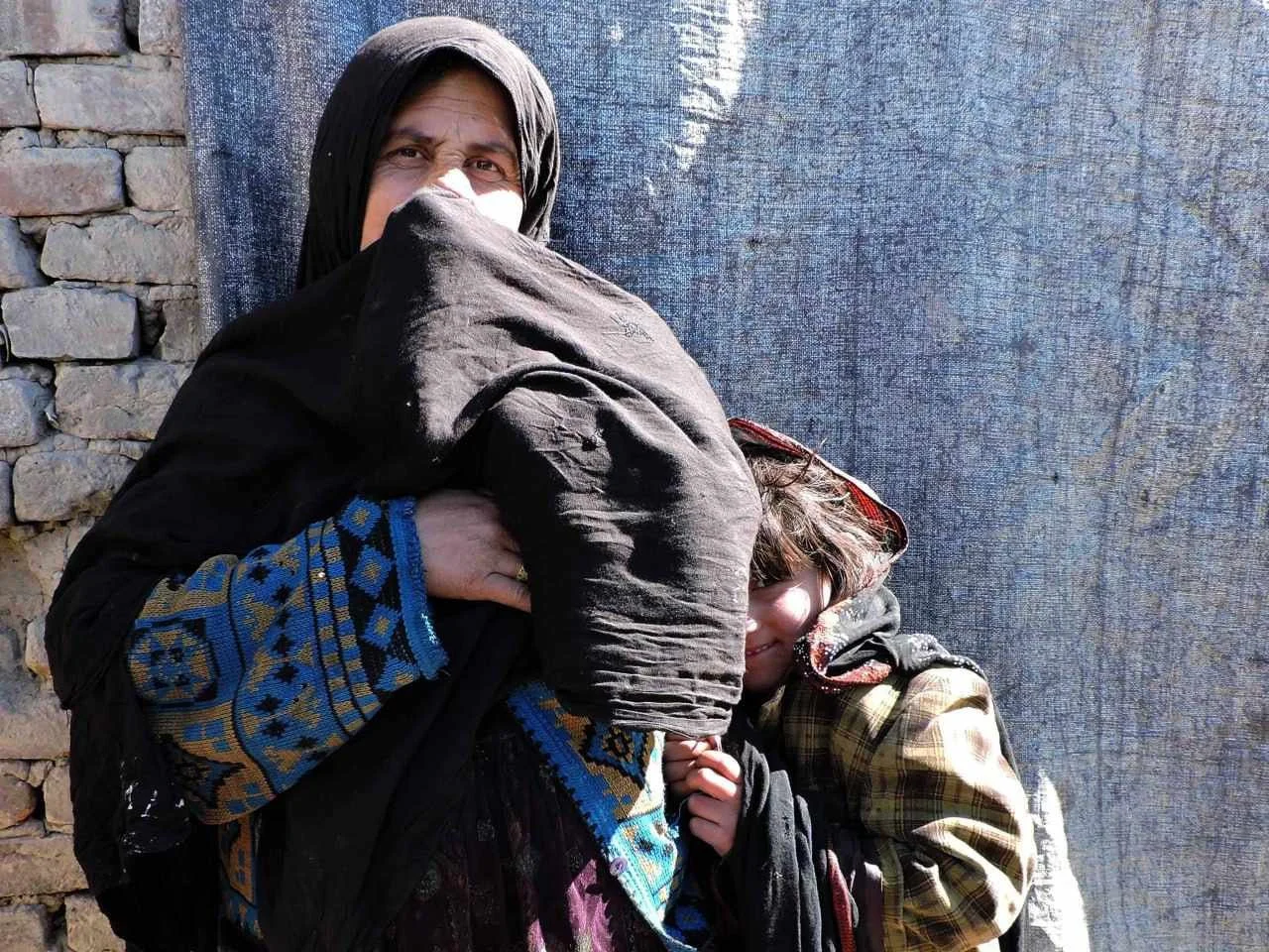 An adult woman and a young child standing outside against a blue fabric backdrop and a brick wall. The woman is holding a black dog with her arm, and the child is smiling and leaning against her.