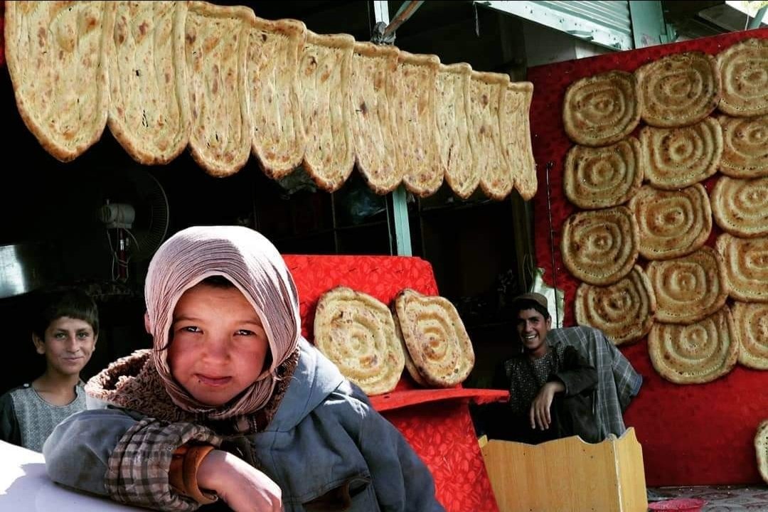 Children standing in front of a display of large, spiral-shaped baked goods, possibly pastries or bread, with some on a red backdrop and others hanging overhead.