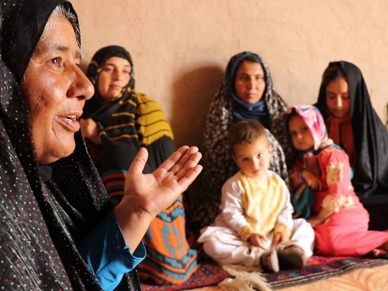 Group of women and children sitting on a carpet, with an elderly woman in the foreground speaking or explaining something.
