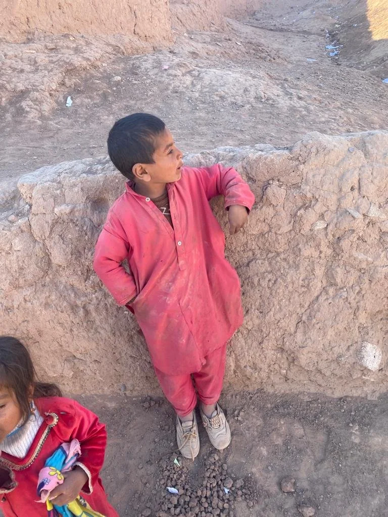 A young boy with short dark hair wearing a dusty red traditional outfit and sneakers, leaning against a dirt wall in a dry, dusty outdoor setting. A girl in a red jacket is partially visible in the lower-left corner of the image.