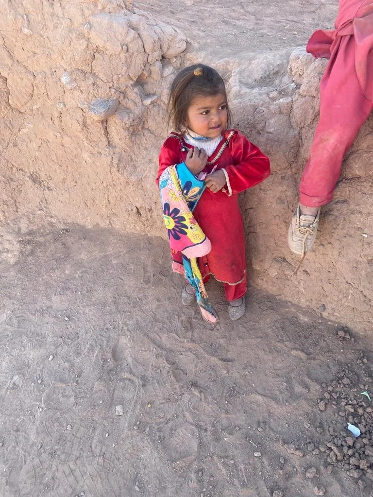 A young girl with dark brown hair in a ponytail, wearing a red dress and colorful blanket, standing on dirt ground beside a dirt wall, with an adult's leg visible nearby.