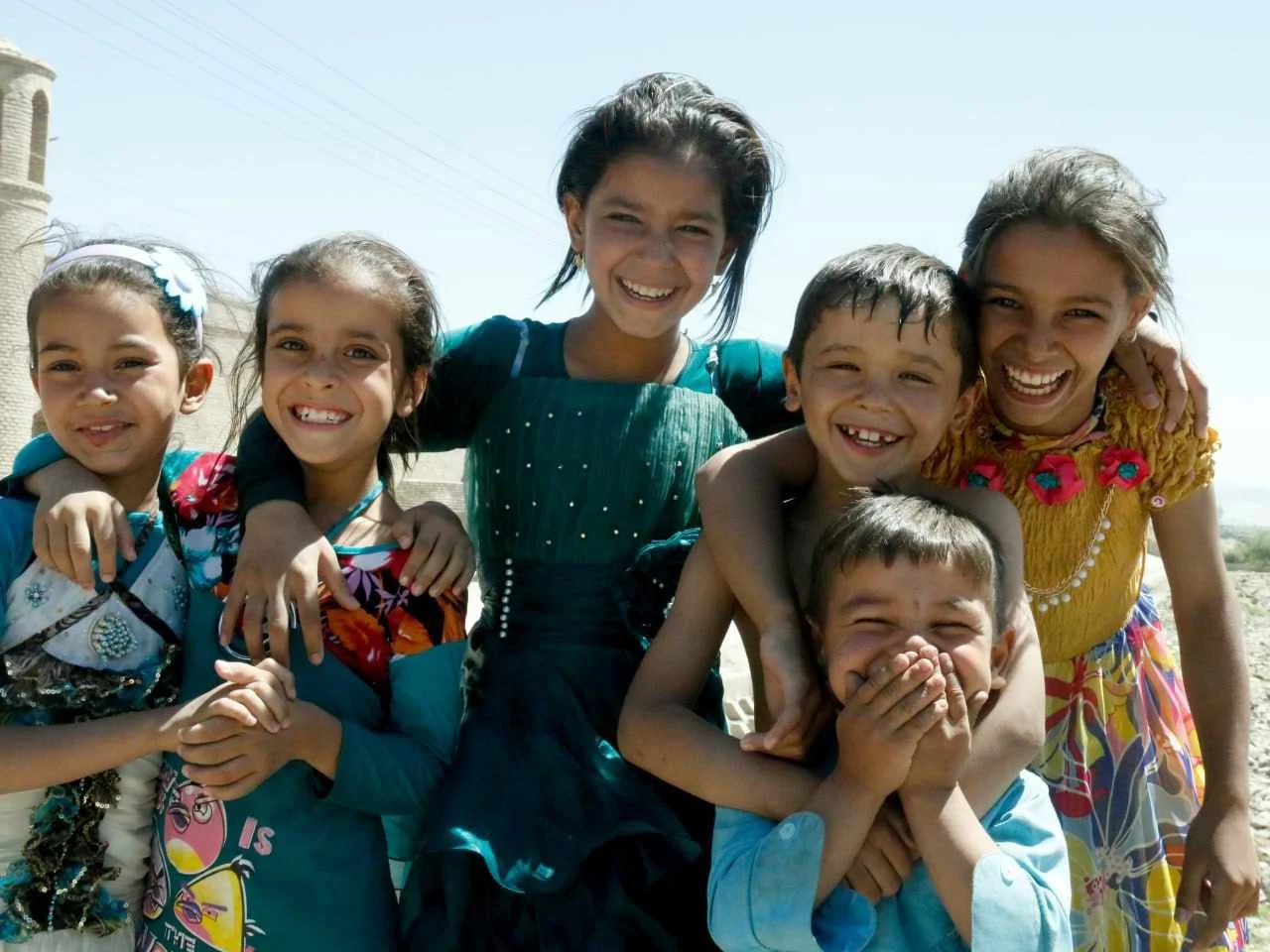 Group of seven children smiling and hugging each other outdoors on a sunny day, with a clear blue sky.