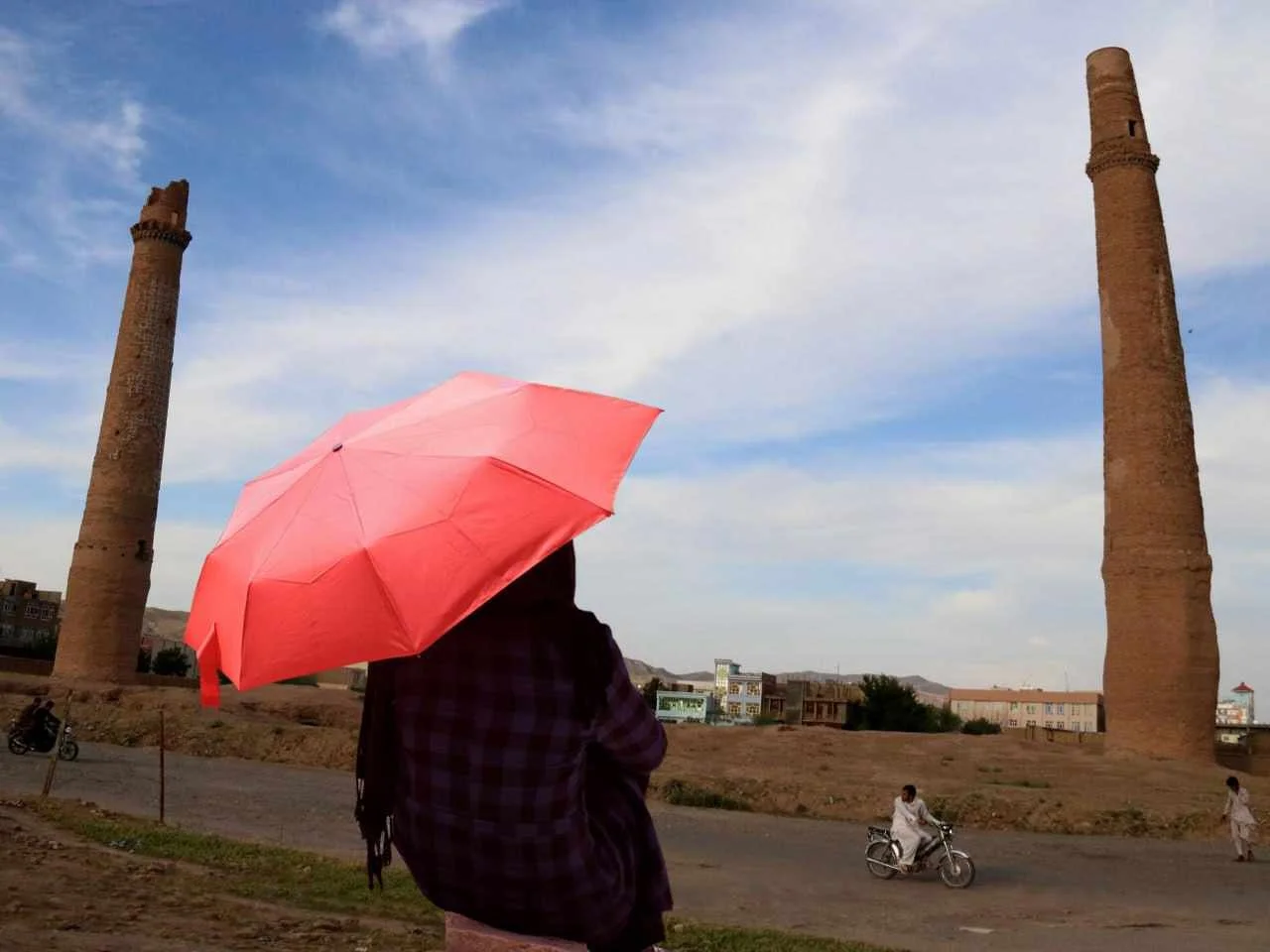 A person holding a pink umbrella stands in front of two ancient brick towers in a rural area, with a person riding a motorcycle and a few others walking nearby under a partly cloudy sky.