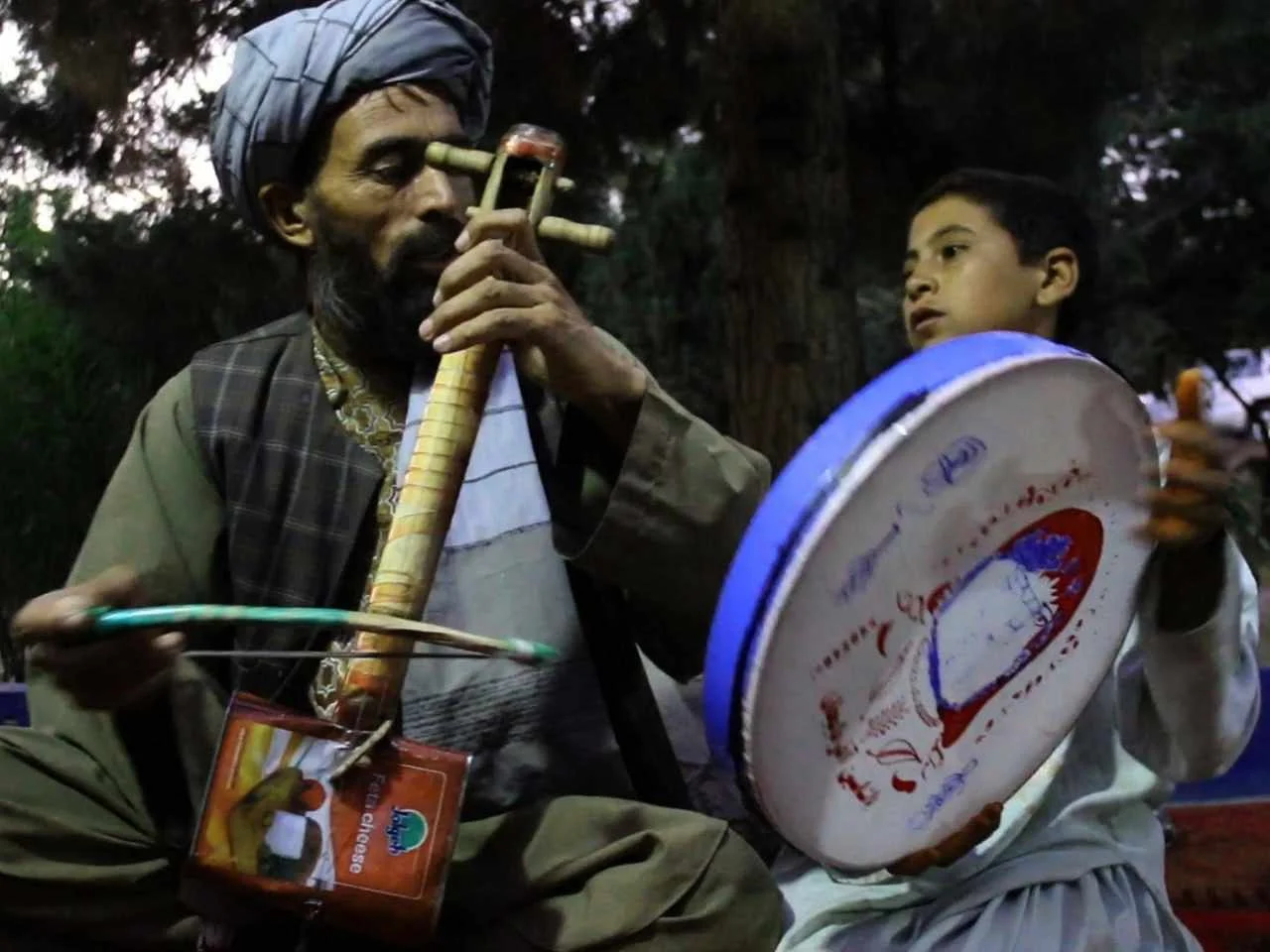 An elderly man in traditional clothing and a turban playing a flute with a young boy holding a large drum, both outdoors with trees in the background.