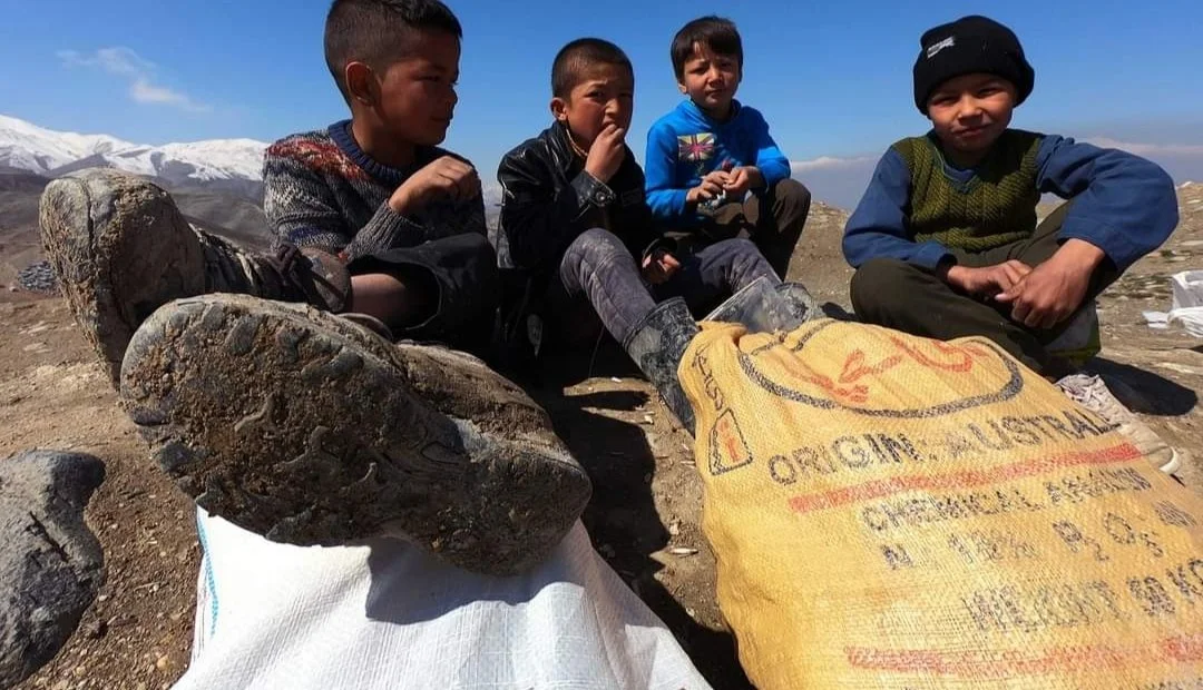 Four children sitting on rocky ground, with snow-capped mountains in the background, one with a bag and rocks in the foreground.