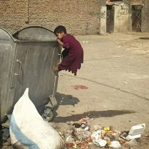 Young boy in purple clothes climbing on a large, open trash bin on a street, with litter and debris on the ground nearby, and a brick wall in the background.