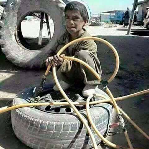 A young boy sitting on a car tire, holding thick yellow electrical cords, in an outdoor area with large tires and vehicles in the background.