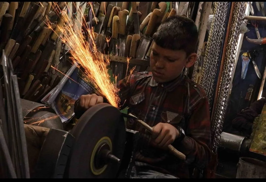 A young boy is grinding metal on a bench grinder, creating sparks in a workshop filled with tools and chains.