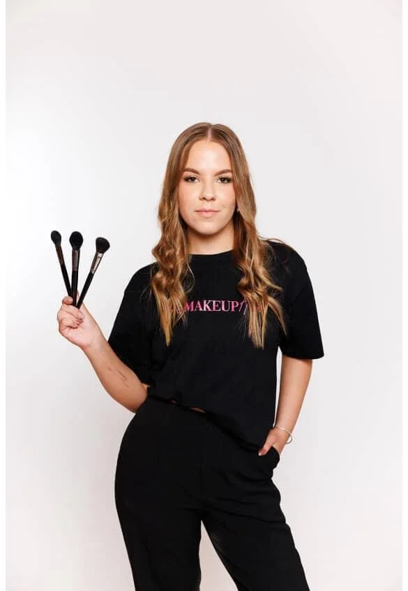 Young woman with long wavy hair holding three makeup brushes, wearing a black t-shirt with pink text and black pants, standing against a plain white background.