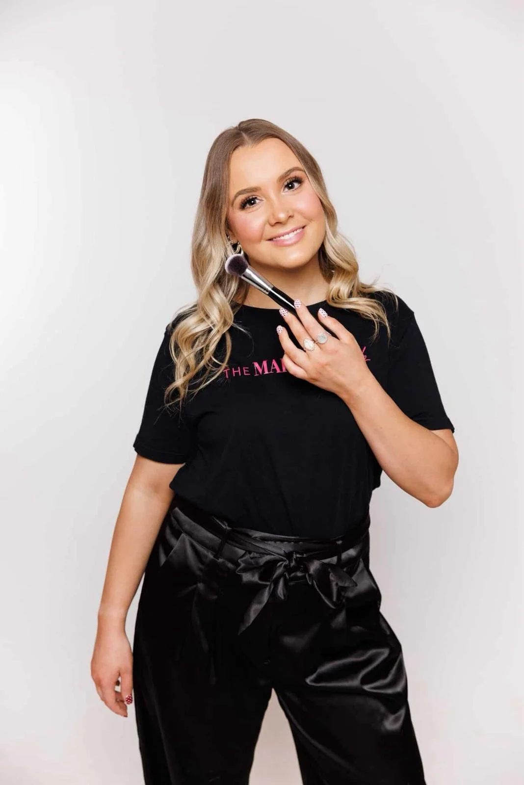 A woman with blonde hair holding a makeup brush to her face, smiling, wearing a black t-shirt and black shiny pants with a bow.