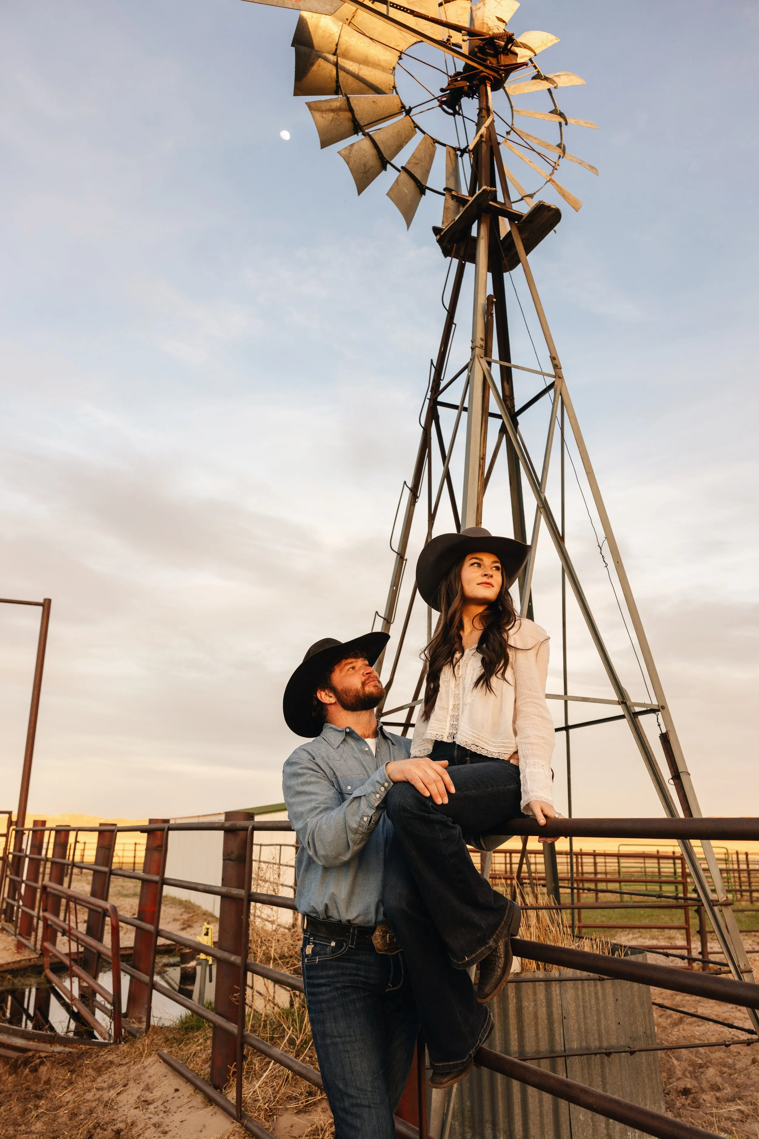 A man in a blue shirt and jeans is holding a woman wearing a black hat and a white top on his lap, standing on a ranch fence with a windmill in the background during sunset.