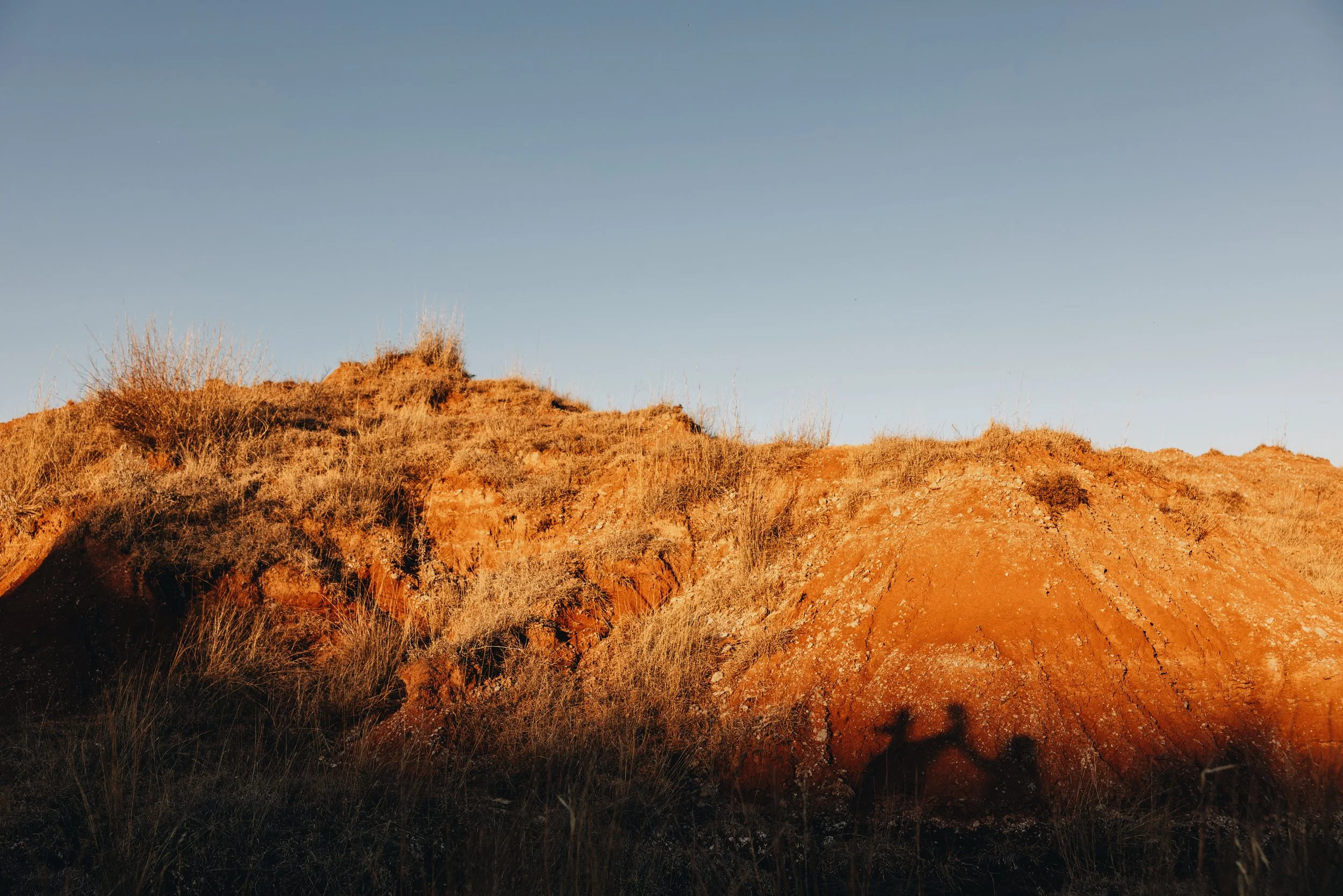 A dry hillside with reddish-brown soil and sparse grass, under a clear blue sky.