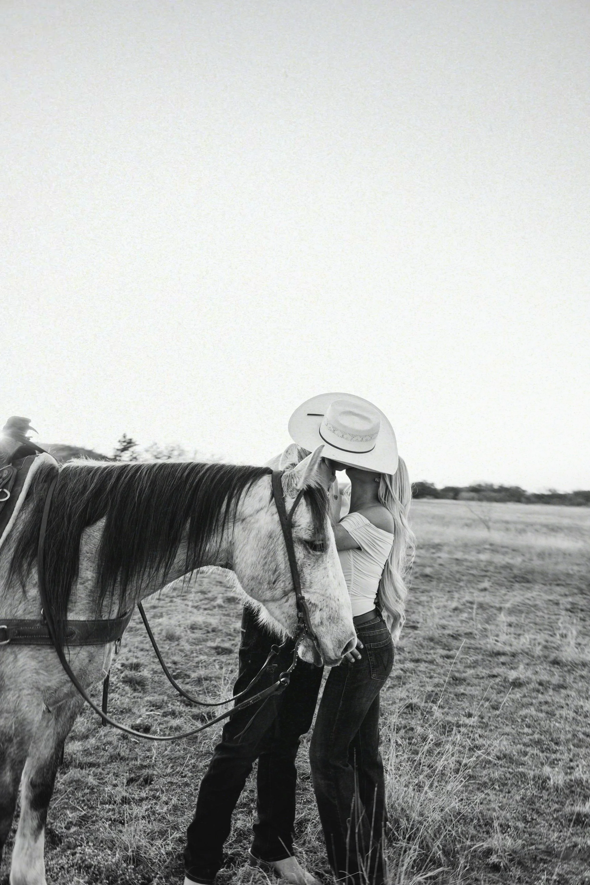 A person standing next to a white and brown horse in a grassy field, wearing a cowboy hat and jeans, with trees in the distance.