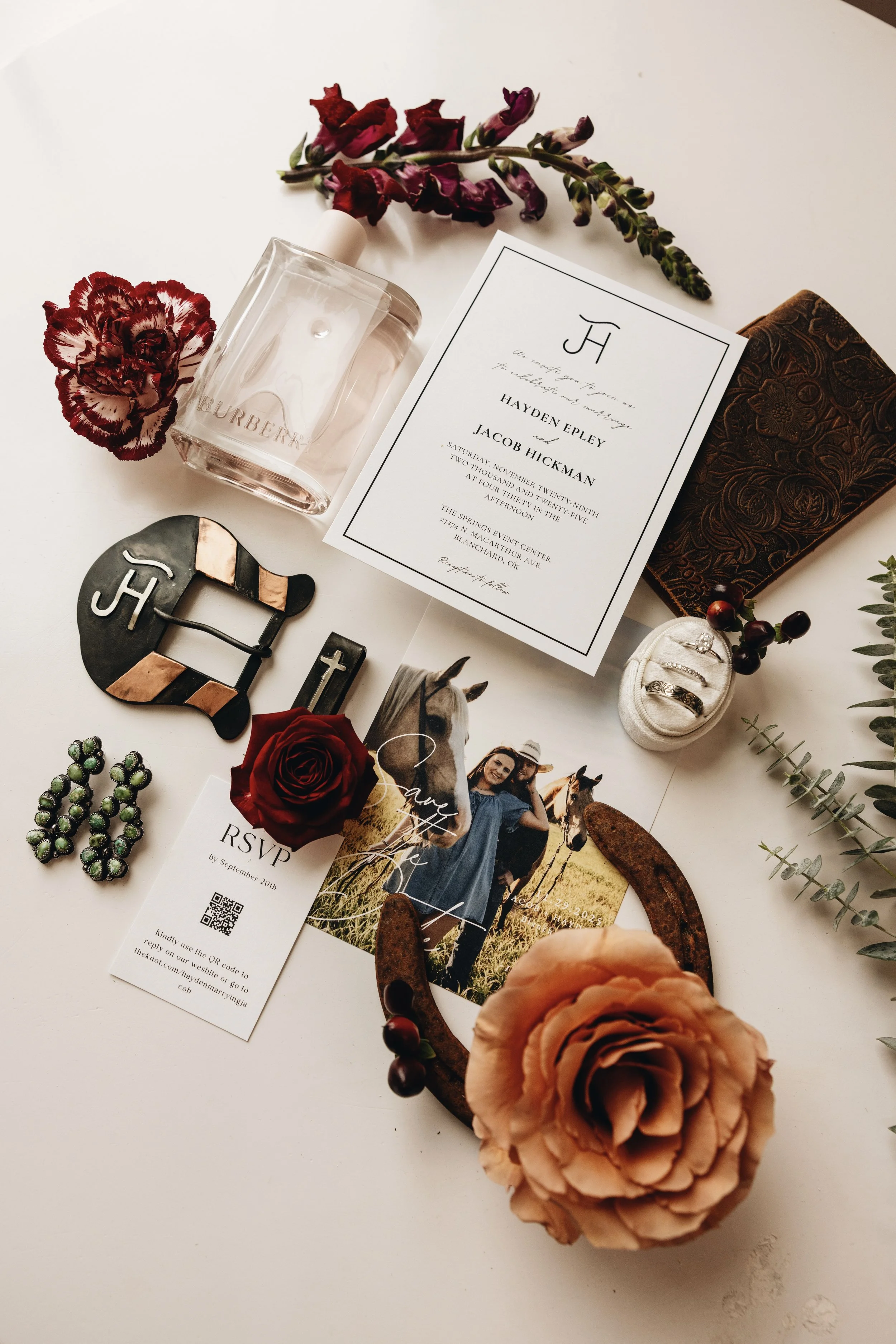 Wedding invitation and accessories including a Burberry perfume bottle, red and purple flowers, a small jewelry box with rings, a decorative belt, wedding photographs, and various wedding-themed items on a white surface.