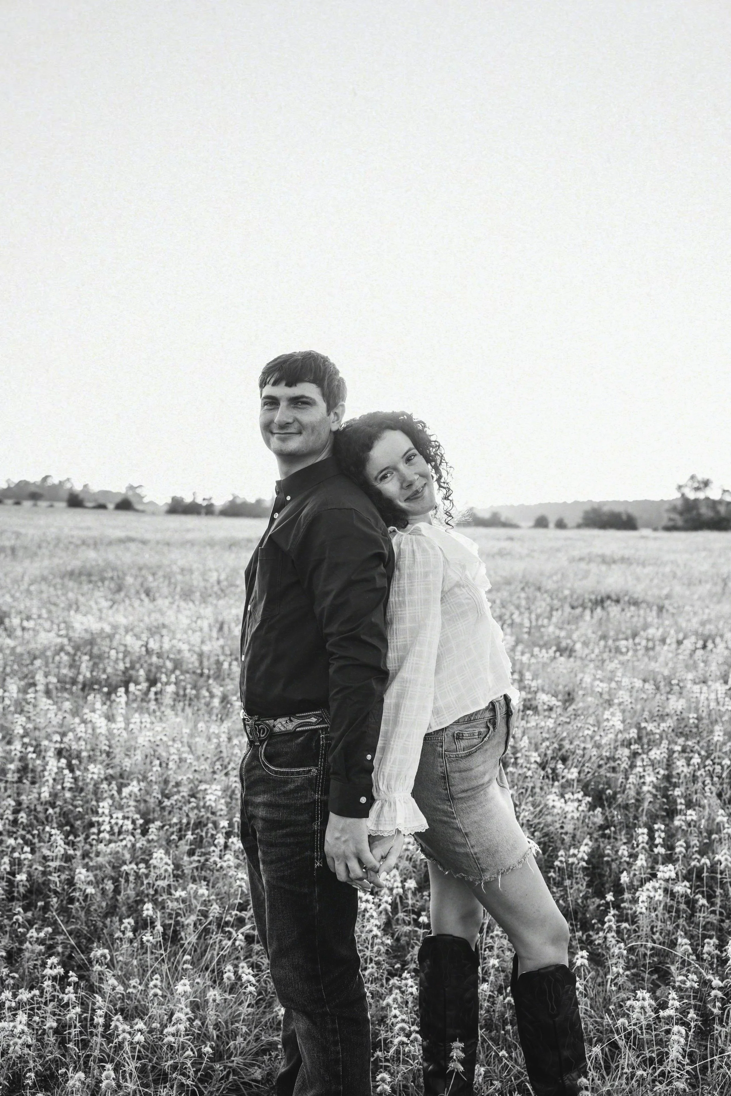 A black-and-white photo of a man and woman standing back to back in a field of flowers, holding hands, with a landscape of trees and open sky in the background.