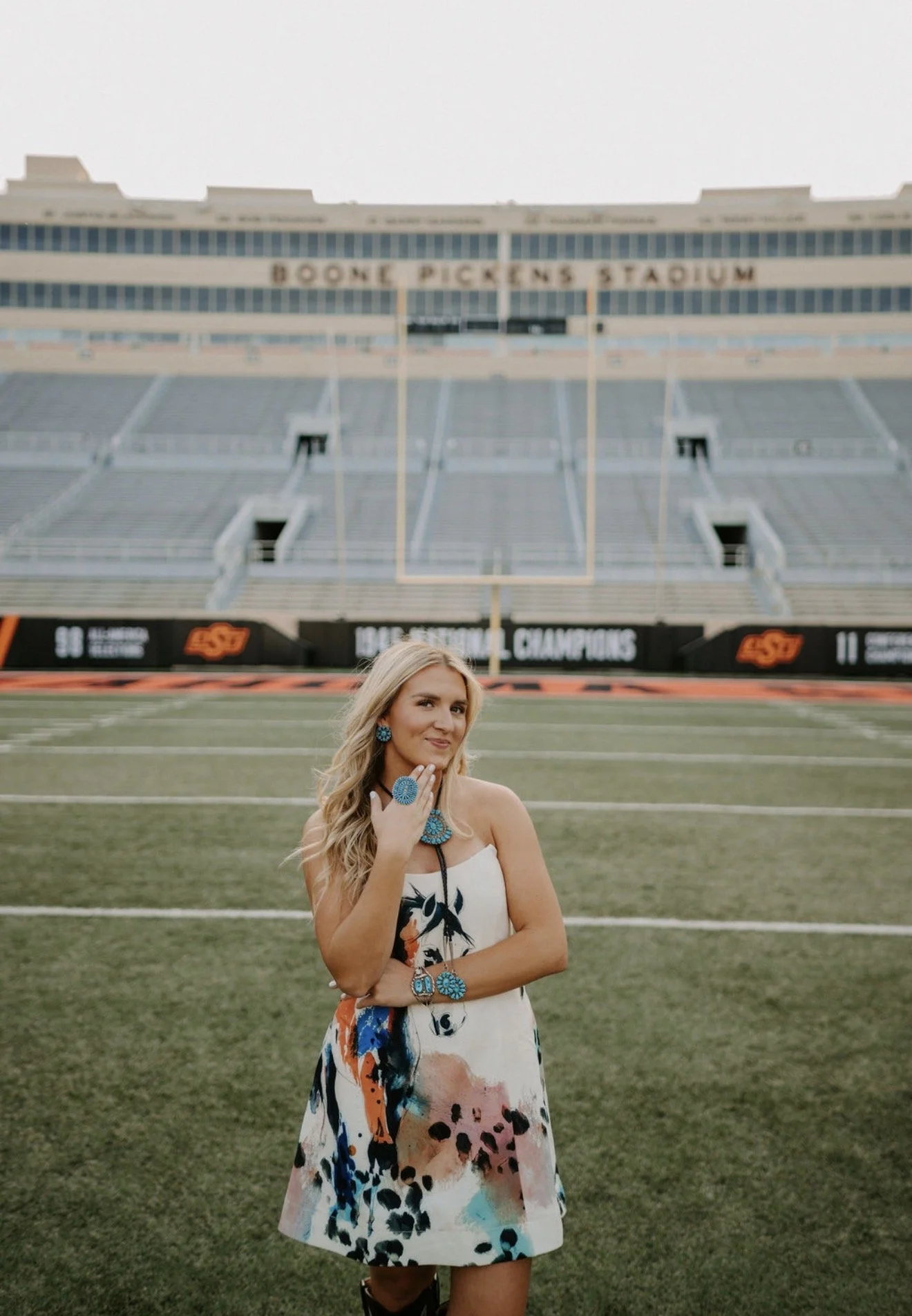 A woman wearing a colorful dress and turquoise jewelry standing on a football field with Boone Pickens Stadium in the background.