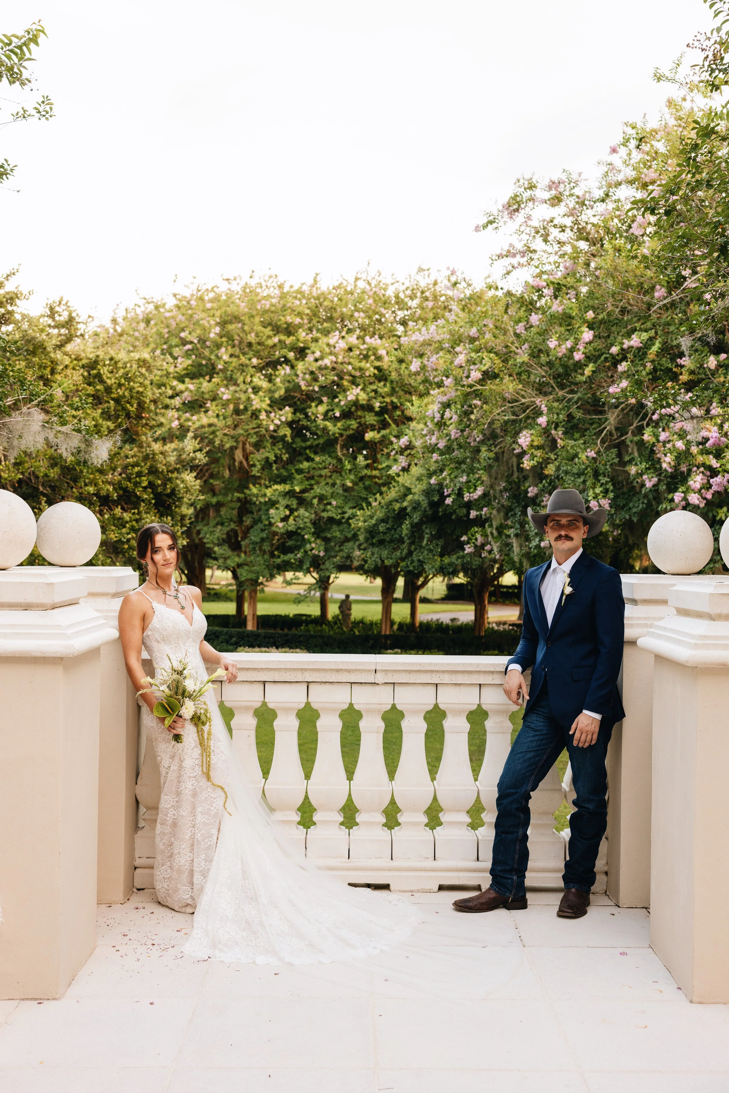 A bride and groom in wedding attire standing on a white balcony outdoors, with large trees and pink flowers in the background.
