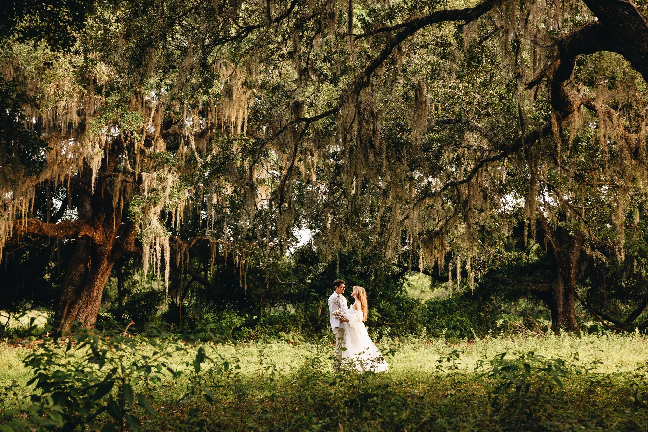A bride and groom standing in a lush outdoor setting under large trees with hanging moss, holding hands and facing each other.