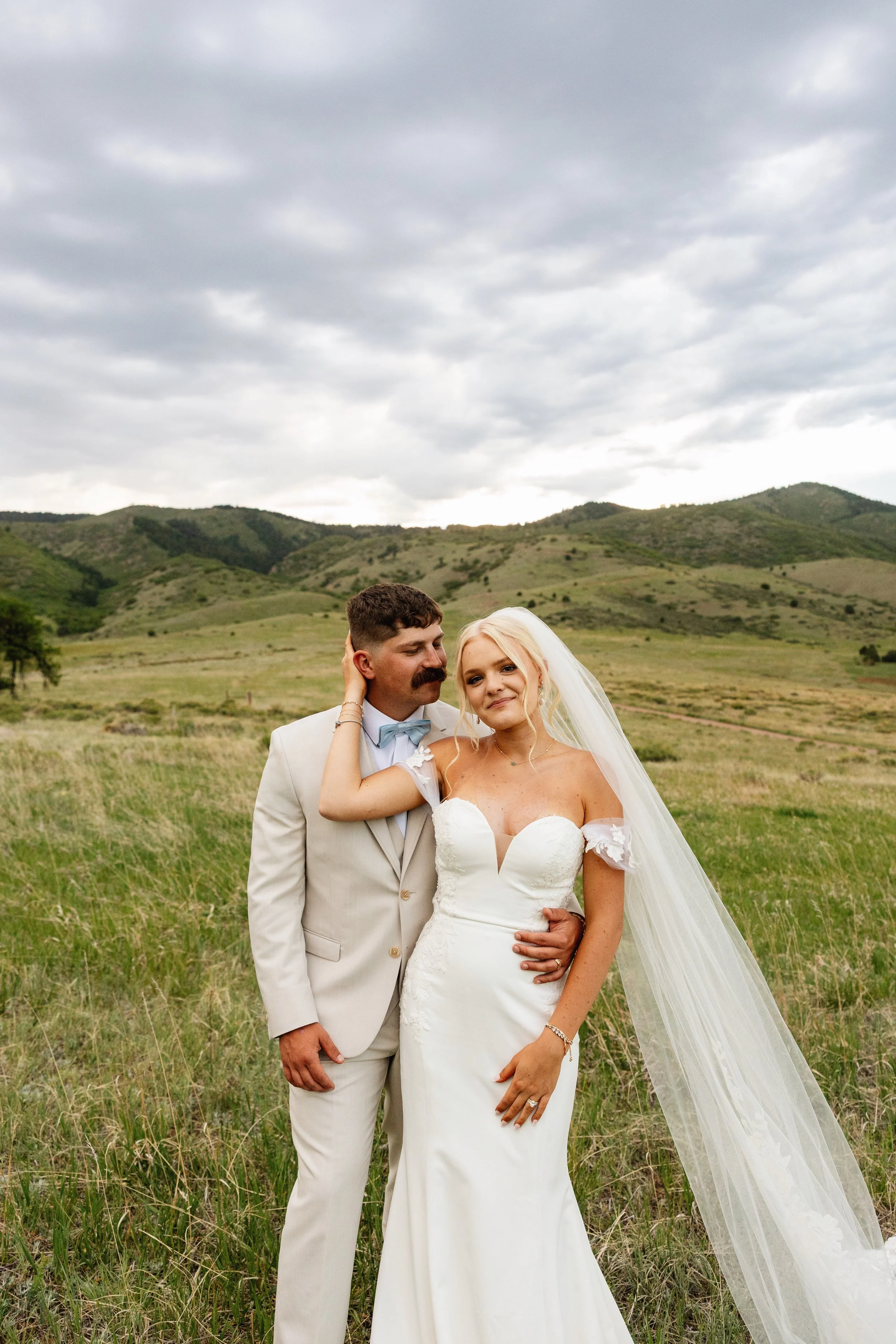 A bride and groom standing together in a grassy field with green hills and a cloudy sky in the background. The groom is dressed in a beige suit with a light blue bow tie, and the bride is wearing a white wedding dress with lace details and a long veil. They are close, with the groom touching the bride's face and the bride smiling softly.