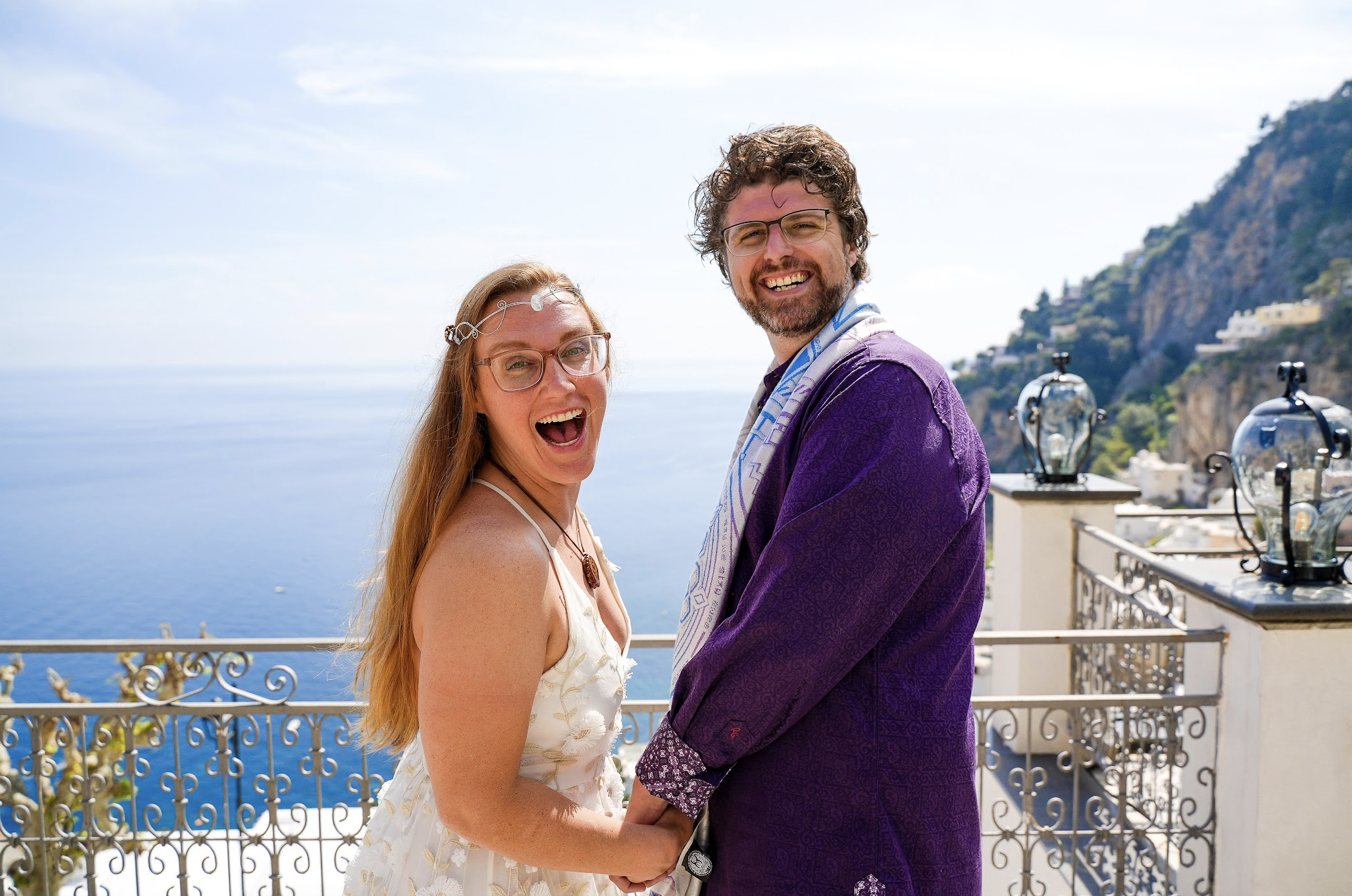 Joyful couple holding hands on a balcony overlooking the ocean with a mountain in the background, sunny weather. Ines Prelovsek wedding photography in Positano, Italy