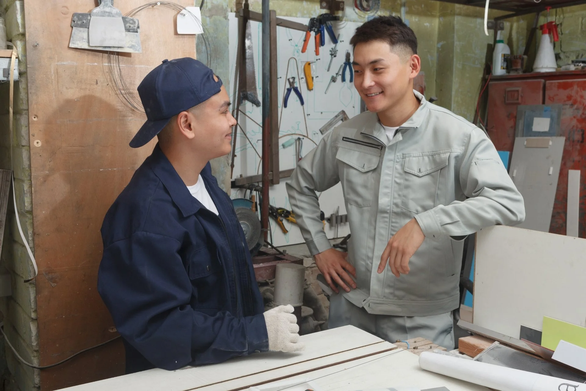 Two young men having a friendly conversation in a workshop. One is wearing a navy cap and jacket, holding a white glove, and the other is in a light gray jacket with hands on hips. Work tools and supplies are visible in the background.