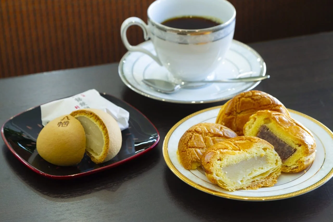 A cup of black coffee on a saucer with a teaspoon, a plate of assorted cream-filled pastries with golden crust, and a steamed bun on a black plate, all set on a dark wooden table.