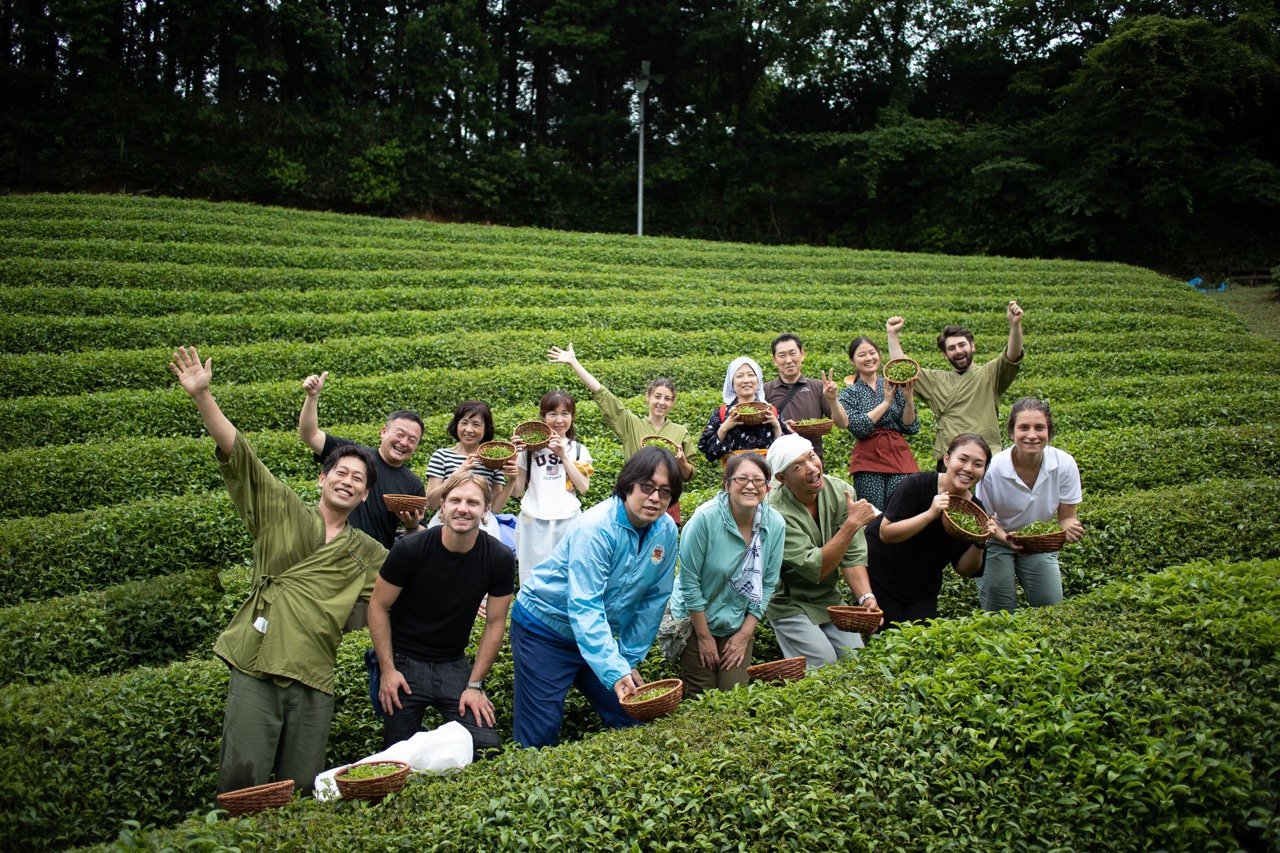 International visitors and local growers smiling together with harvest baskets in the tea fields of Kyoto Obubu Tea Farms in Wazuka.