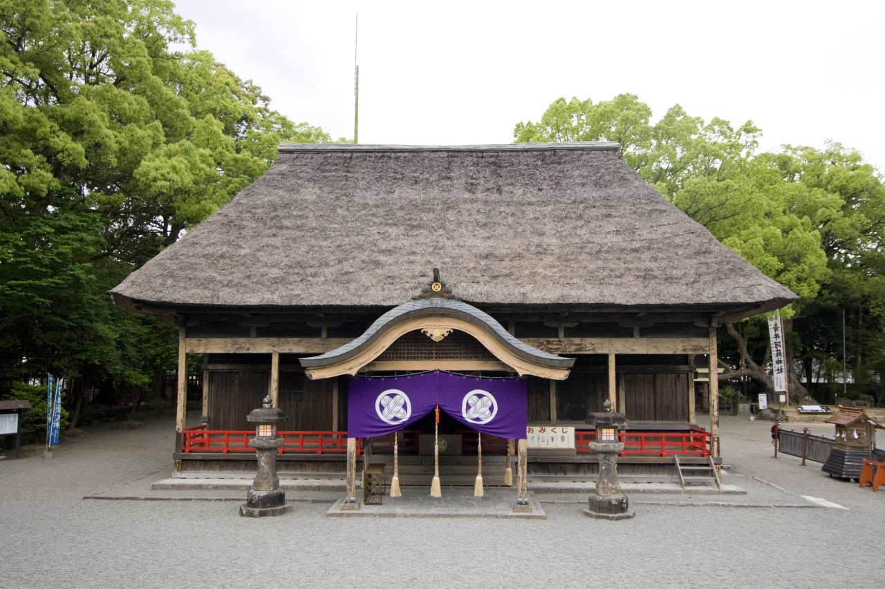 Thatched-roof haiden (worship hall) of Aoiasojinja Shrine in Hitoyoshi, with purple noren and stone lanterns framed by lush green forest — a hidden National Treasure of Kyushu.