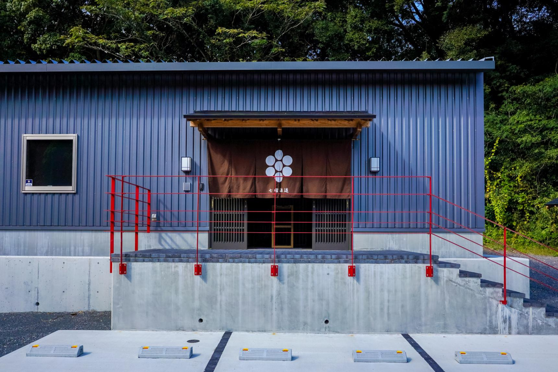 Shichiyo Sake Brewery in Okagaki — modern dark metal facade with a seven-star noren and red railings, framed by lush green trees.