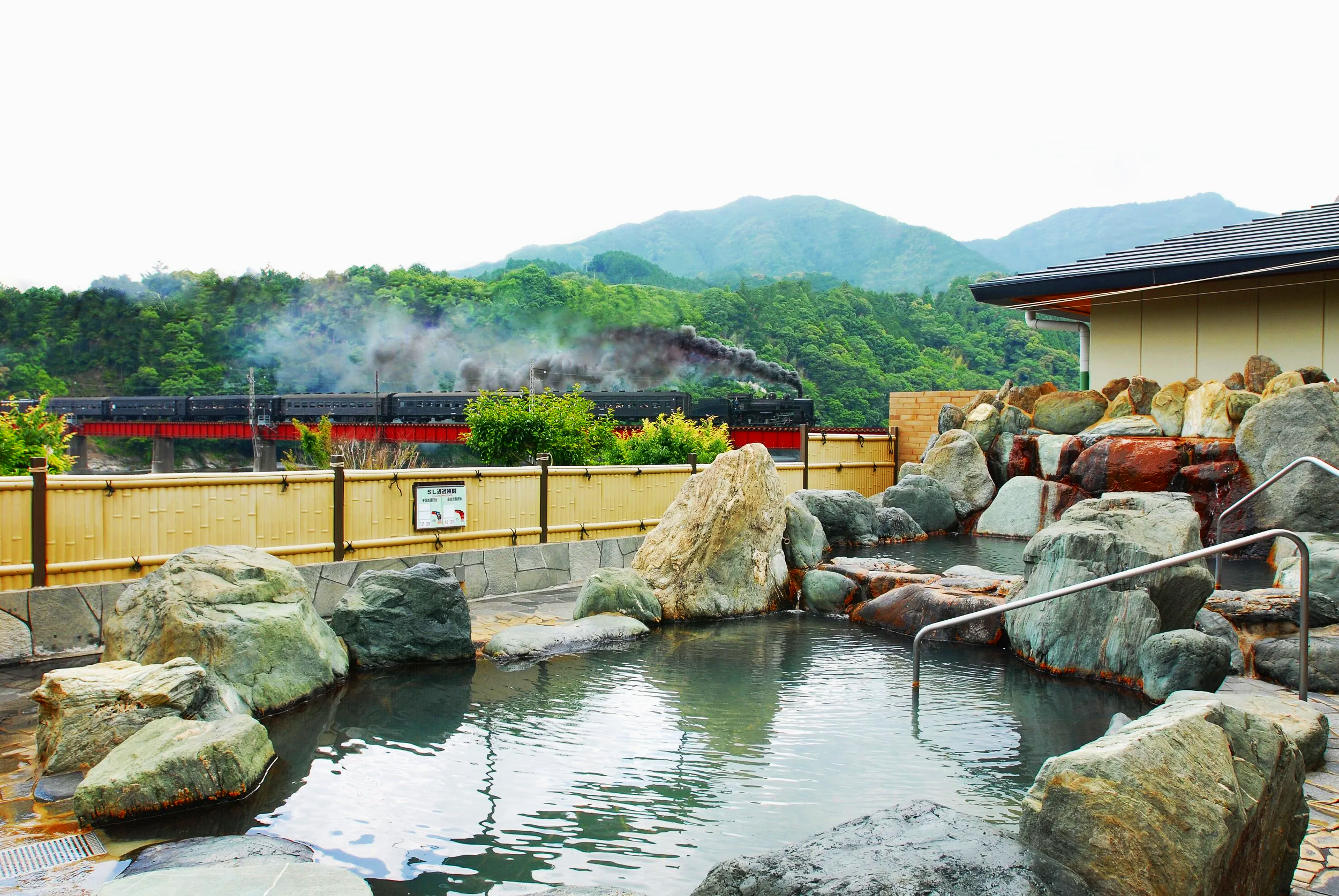 Open-air onsen with rocky bath and steam locomotive crossing a red bridge through misty mountain forests at Kawane Onsen, Shimada.