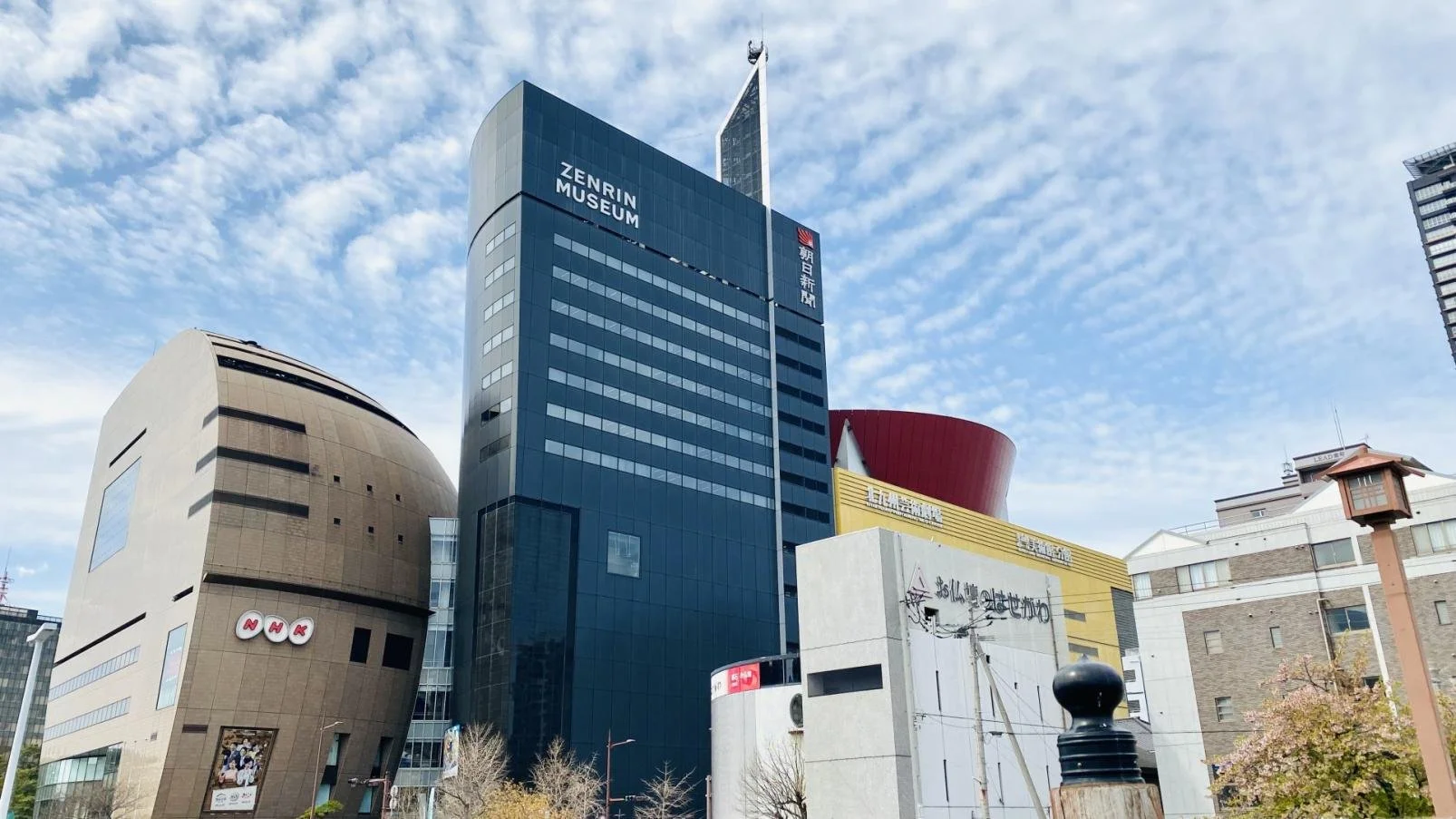 Exterior view of Zenrin Museum, a striking dark-navy high-rise in Kokura, Kitakyushu, surrounded by distinctive local architecture under a bright blue sky.
