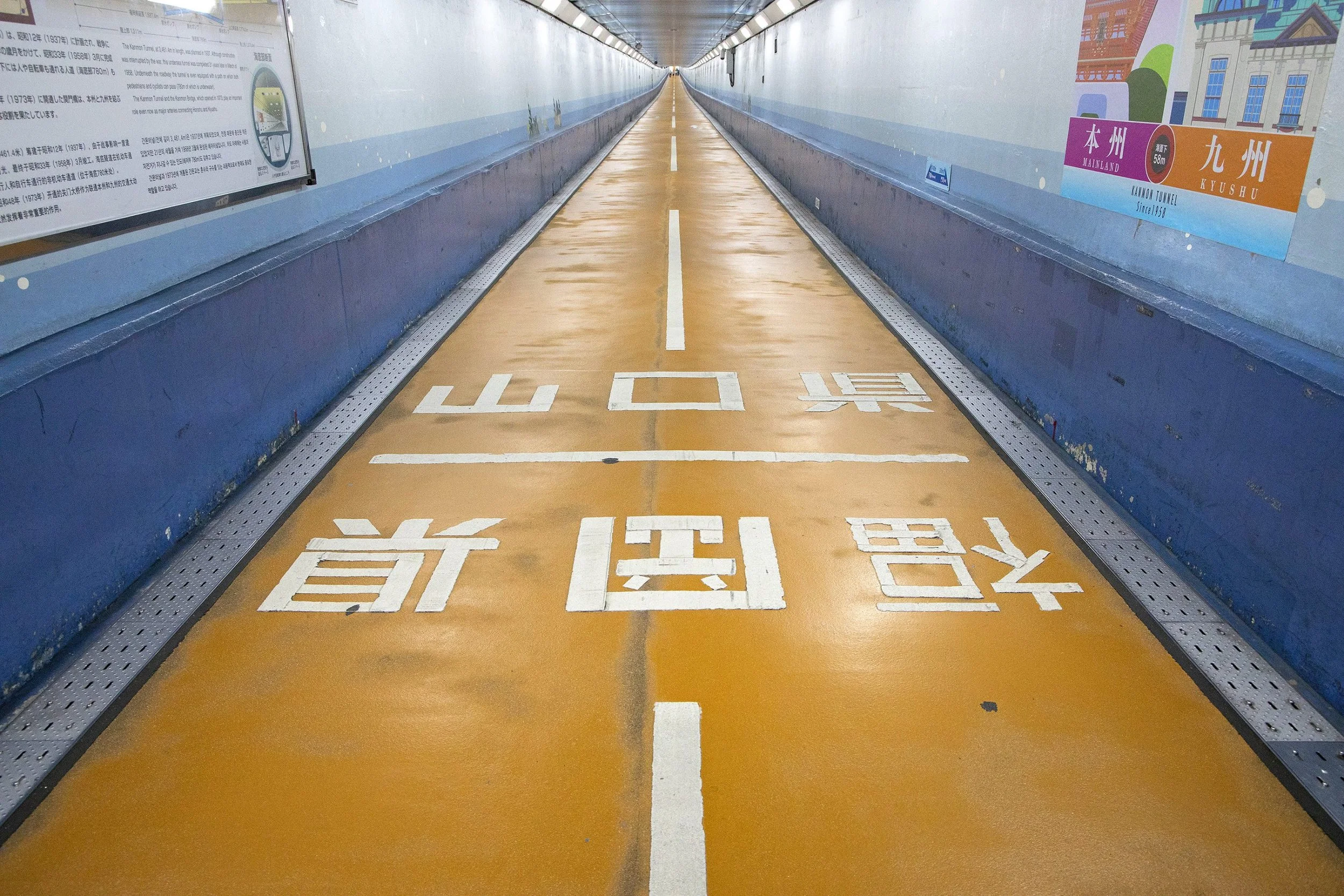 Interior view of the Kanmon Tunnel Footpath, an orange-floored undersea walkway with blue walls connecting Fukuoka and Yamaguchi in Kitakyushu, Japan.