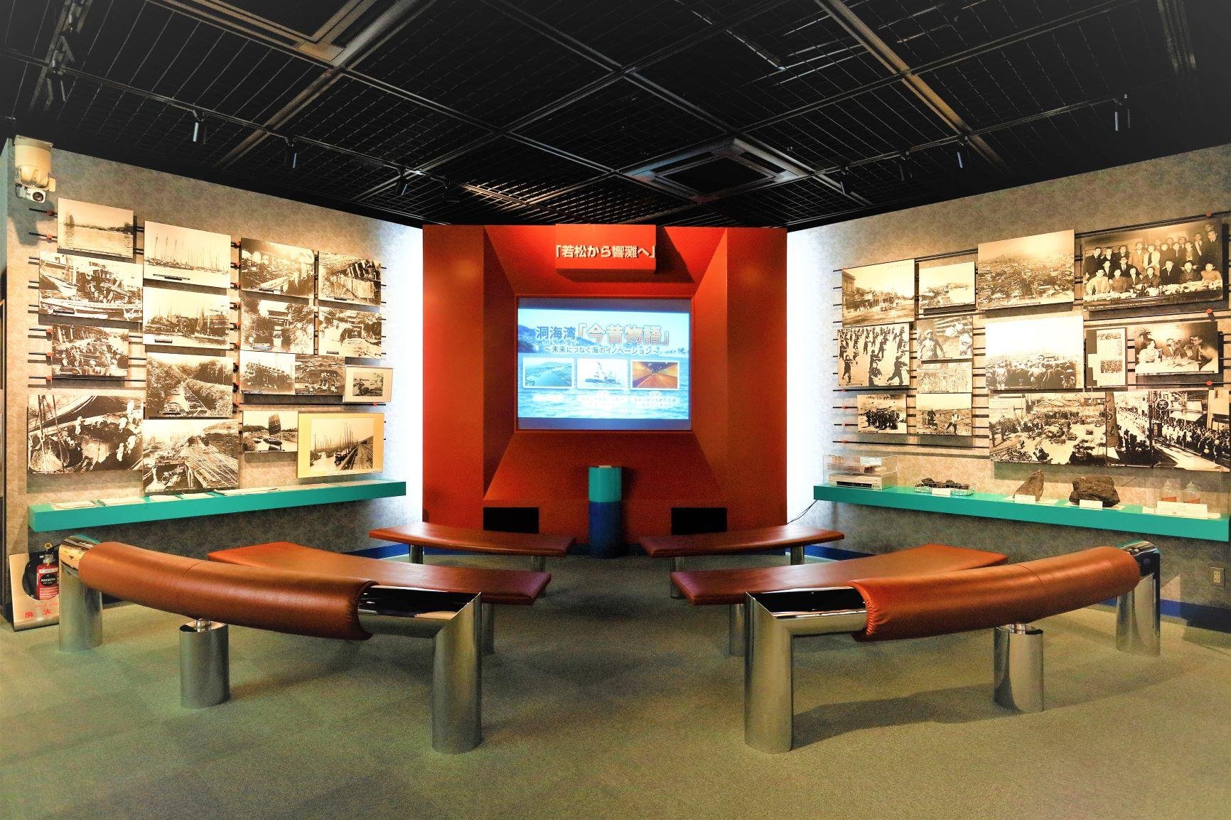 Interior exhibition room at Wakachiku Museum in Kitakyushu, with curved benches facing a screen and walls of black-and-white photos tracing Dokai Bay's coal-port history.