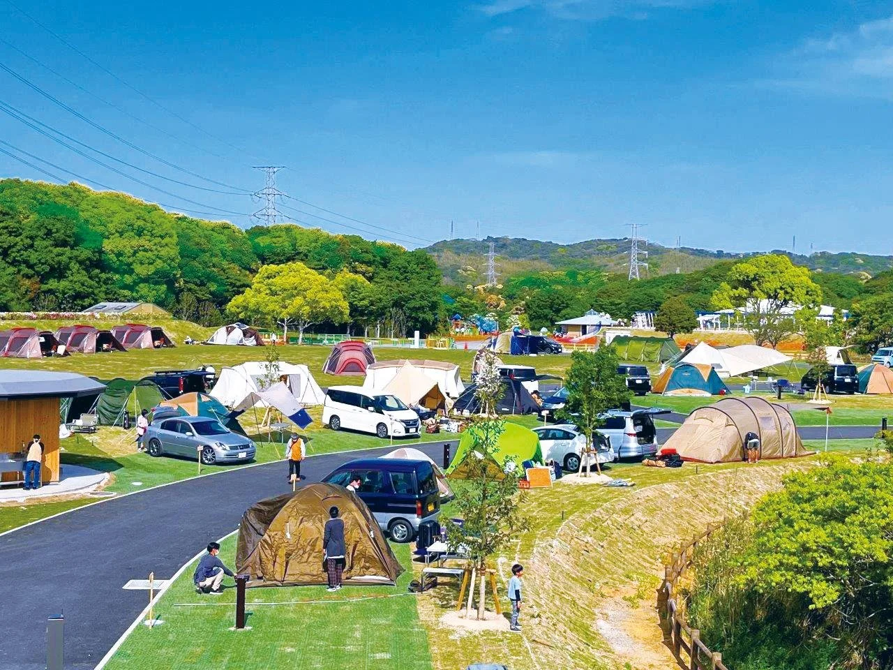 Colorful tents and family campers spread across the sunny green lawns of HIBIKINADA CAMP BASE in Kitakyushu, framed by wooded hills.