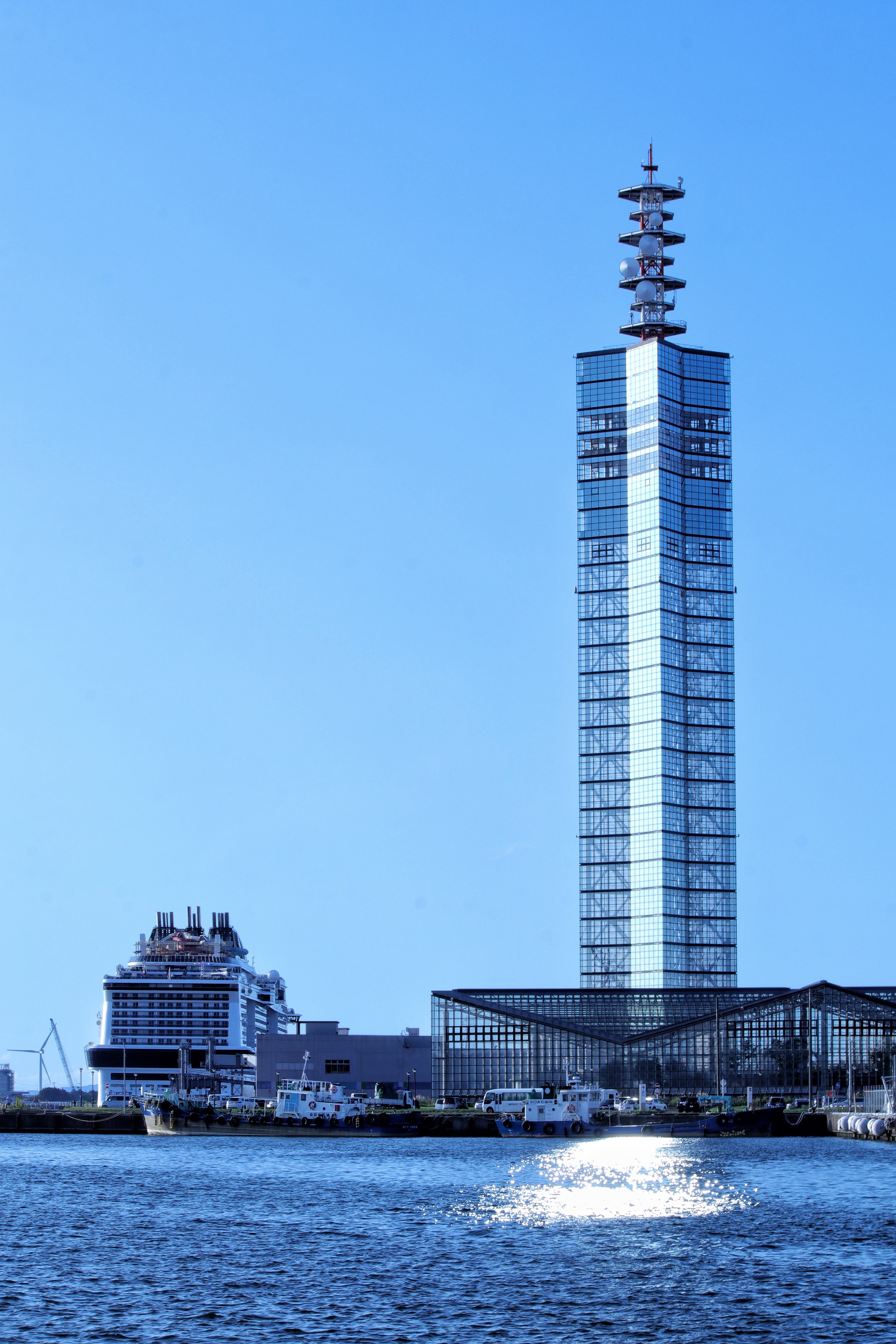 Glass Port Tower Selion rising against a clear blue sky above Akita Port, with a cruise ship and boats docked at the sunlit waterfront.