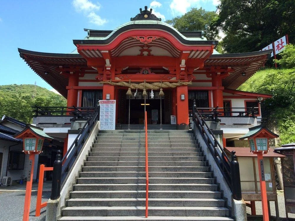 Vivid vermilion main hall of Awashima Shrine in Moji, Kitakyushu, framed by stone stairs, green forest, and clear blue sky — a peaceful 850-year-old sanctuary.